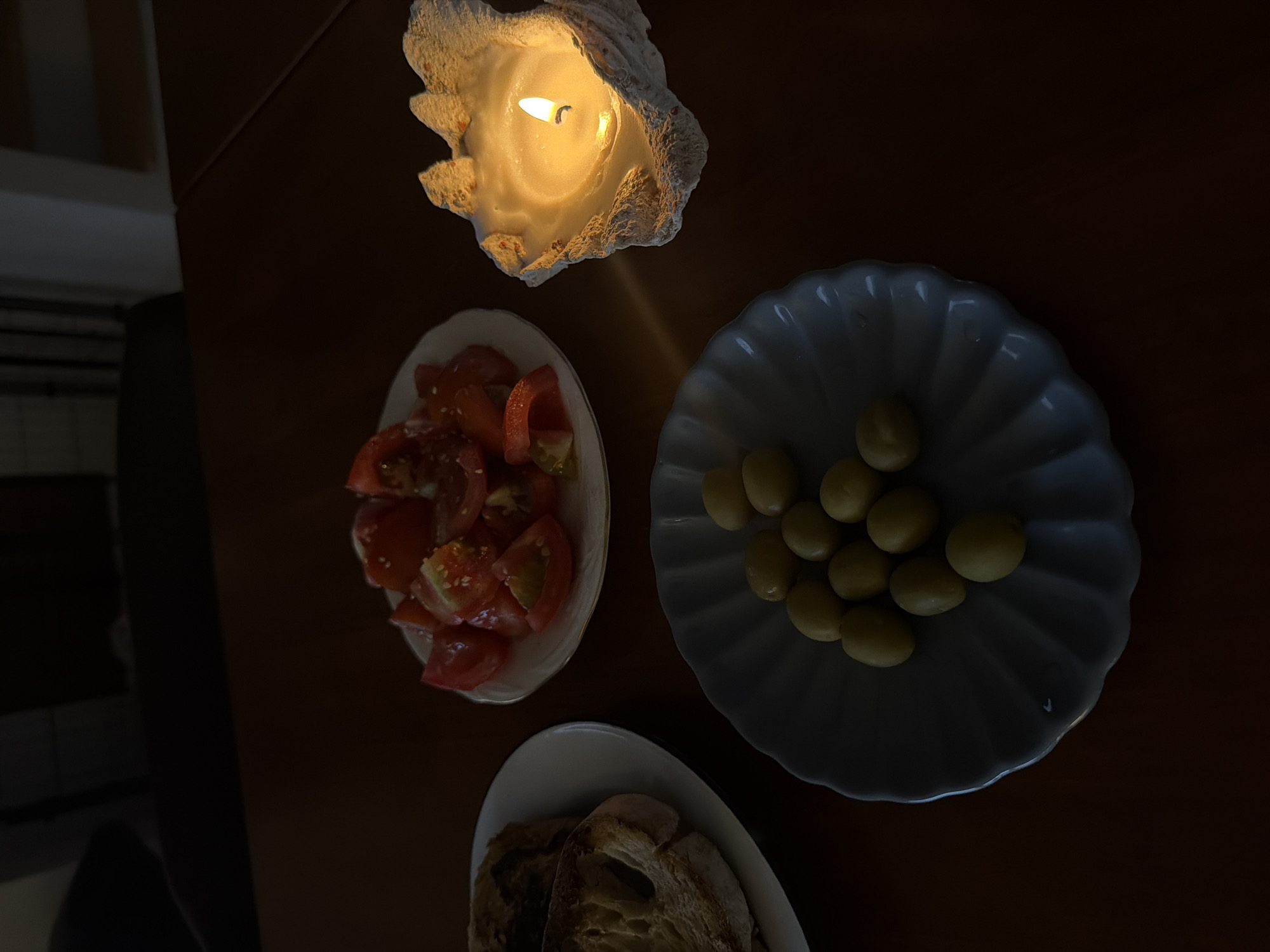 A dimly lit scene presents a dark brown wooden table illuminated by a candle. A candle encased in a decorative, possibly shell-like, holder sits at the top of the frame. Three white plates are arranged around the candle. The first holds red tomato slices seasoned with sesame seeds. The second is piled with olives. The third plate, at the bottom of the frame, has slices of toast. It is unknown if the scene is in Chang Phueak, Thailand. No people or interactions are visible in the photo.