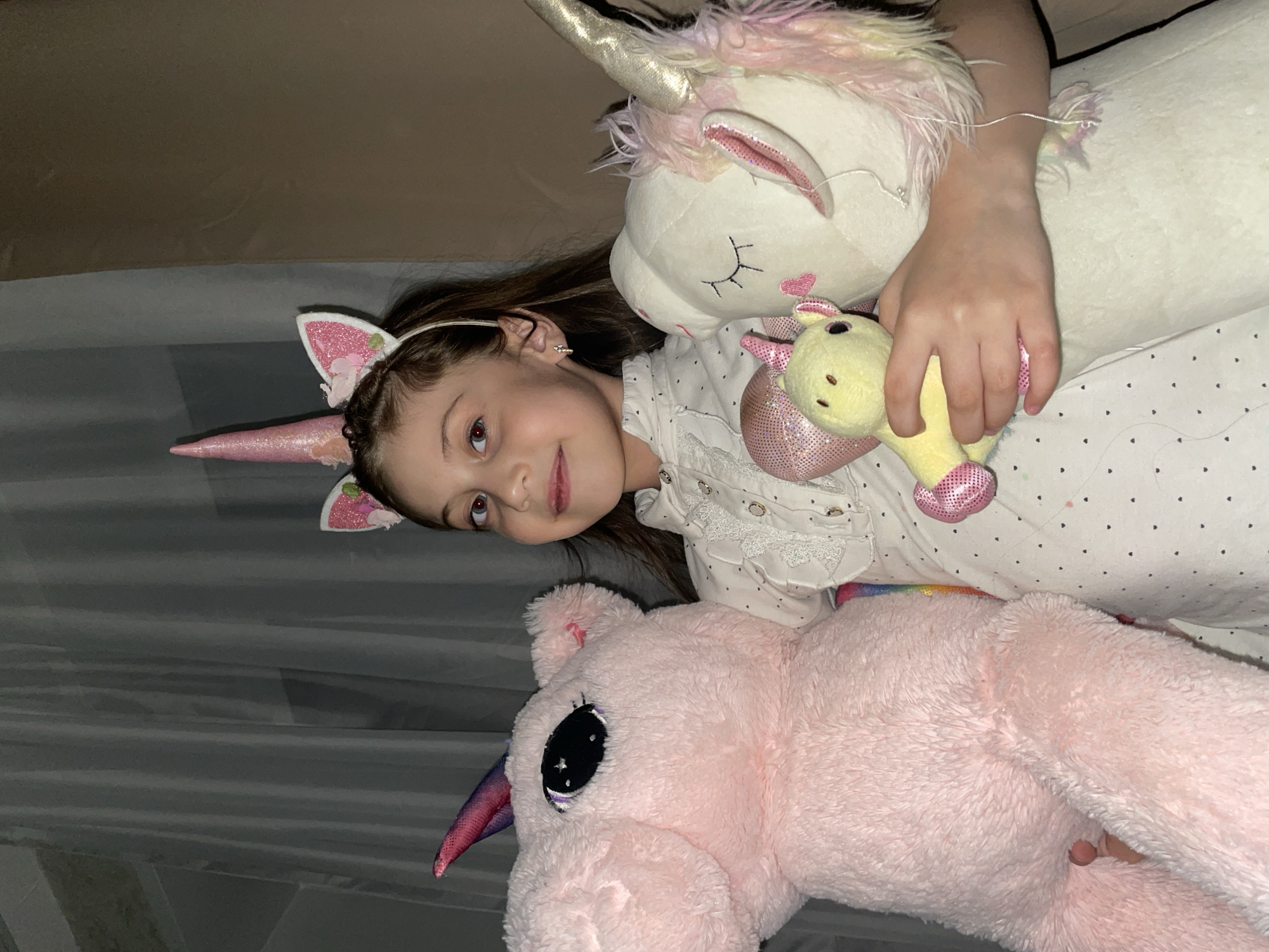 A young girl wearing a unicorn horn headband and earrings is lying down and smiling. She is surrounded by three stuffed unicorn toys: a large pink one, a large white one, and a small yellow one. The girl is wearing a white top with a polka dot pattern. The background is blurry and indistinct. There is no discernible evidence to indicate this image was taken in Izhevsk, Russia.