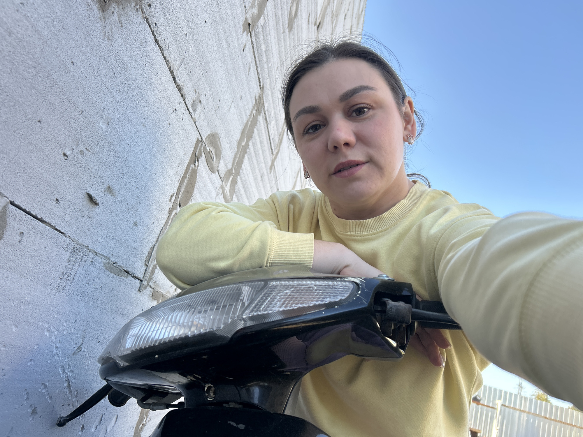 A woman with dark hair is posing for a selfie on a scooter. She is wearing a yellow sweater. She is leaning against a white cinder block wall. The sky is blue. There is a white fence visible in the background. The location is likely in Chaikovsky, Russia, based on the instructions.