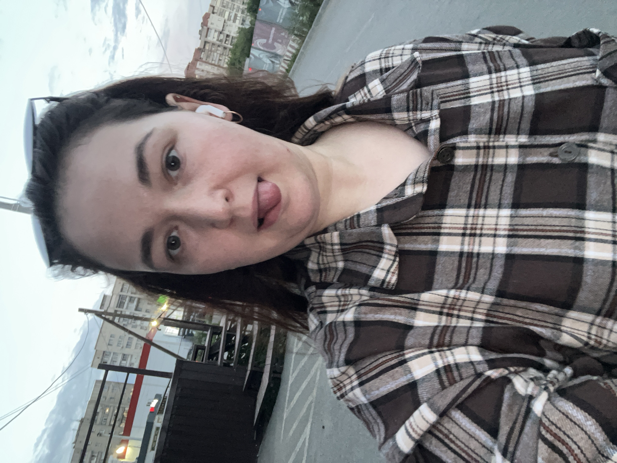 The image is a slightly distorted, low-angle selfie of a young woman. She has dark hair and is wearing a brown, white, and black plaid shirt. She is sticking her tongue out slightly. In her ears, she has white wireless earbuds. Sunglasses are pushed up on top of her head. The background shows a cloudy sky and buildings, suggesting an urban setting. There are also some metal railings and what appears to be a dark, rectangular dumpster or trash bin in the foreground. The lighting suggests it might be late afternoon or early evening. The image appears to have been taken outdoors, possibly in a public area like a street or park.