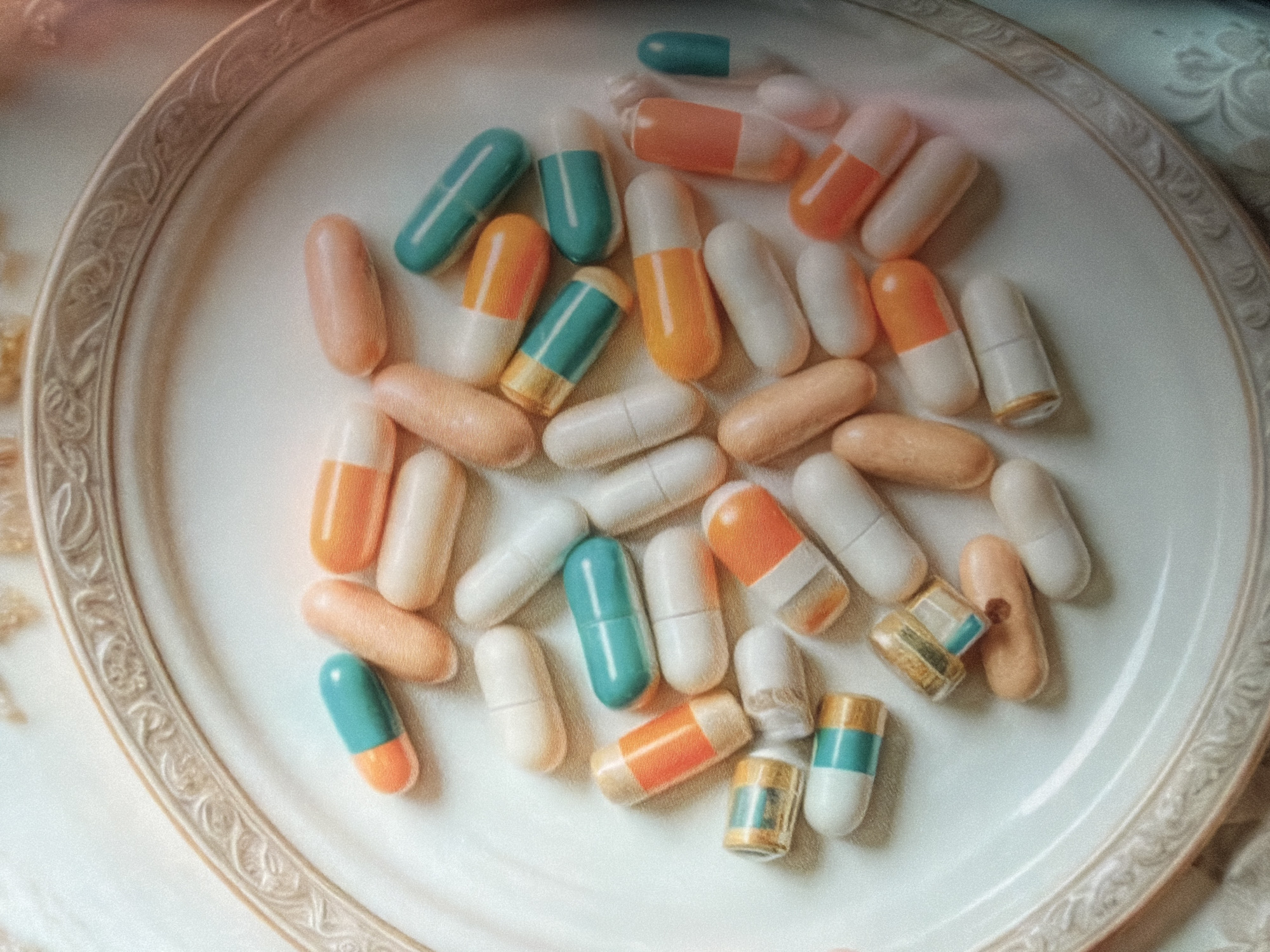 The image shows an assortment of capsules on a decorative white plate with an ornate, raised rim. The capsules are of varying colors, including combinations of white, peach, orange, and teal. Several capsules appear to be split open, with some showing their contents. The setting seems to be indoors, possibly on a table covered with a fabric that has a subtle pattern. The image does not contain sufficient information to determine its location in the city of Moscow, Russia, or provide any additional details about the scene.