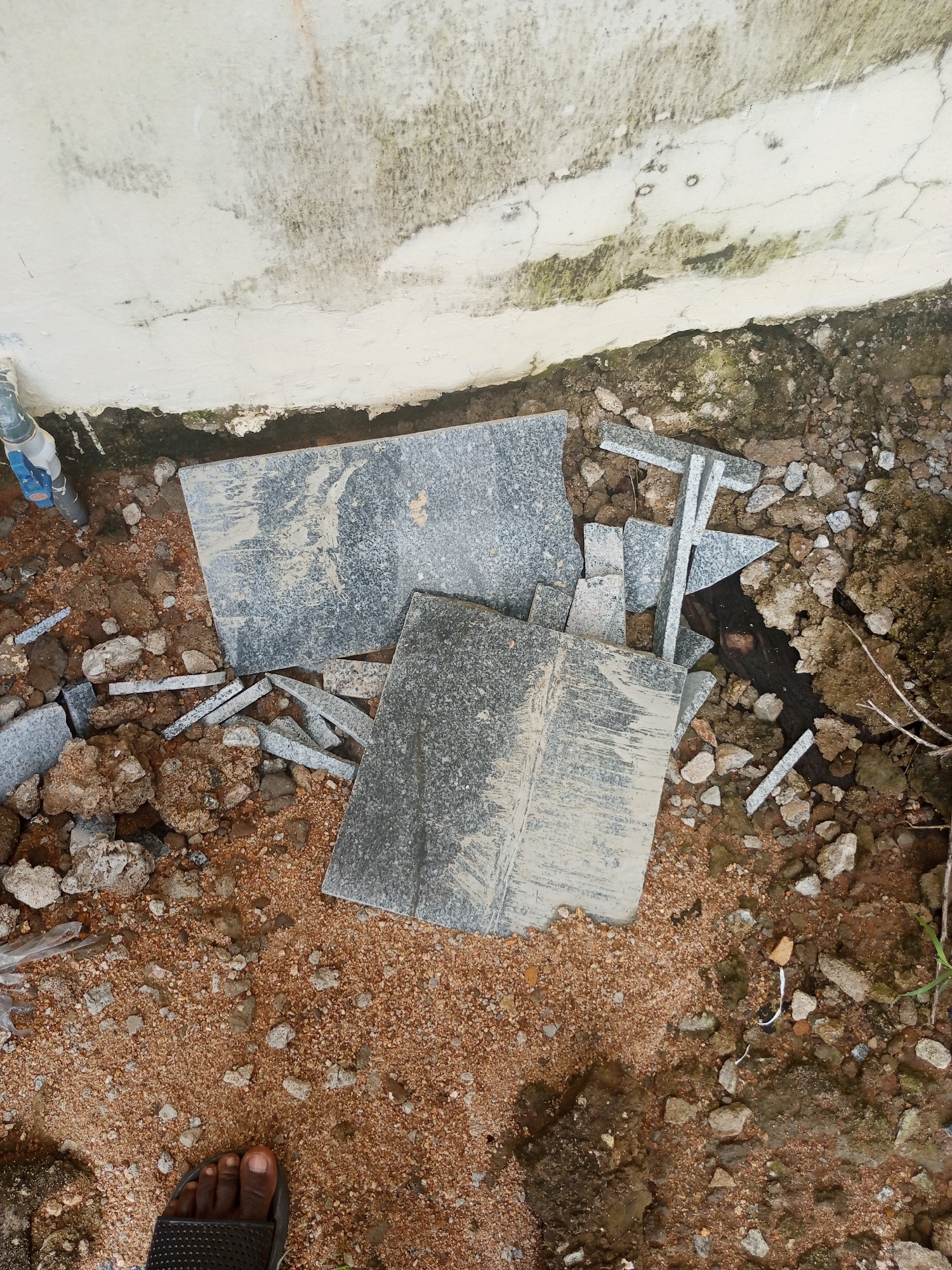 The image depicts a pile of broken granite tiles scattered on the ground, surrounded by reddish-brown sand and soil. The tiles are various sizes, with some larger pieces and many smaller fragments. The granite has a speckled pattern of black and white. In the background, there is a light-colored, weathered wall with some green and black staining, possibly indicating dampness or algae growth. A section of a pipe, possibly a water pipe, with a blue component, is visible on the left side of the image, emerging from the ground near the wall. The ground also contains various rocks and pebbles. In the lower left corner, a person's left foot wearing a black sandal is visible, standing on the sandy ground. The overall scene suggests an outdoor construction or renovation area, possibly a backyard or an unfinished building site. There is no clear indication of the time of day or weather, but the lighting suggests it is daytime. No visible text is present in the image. The location context is Jalingo, Nigeria.