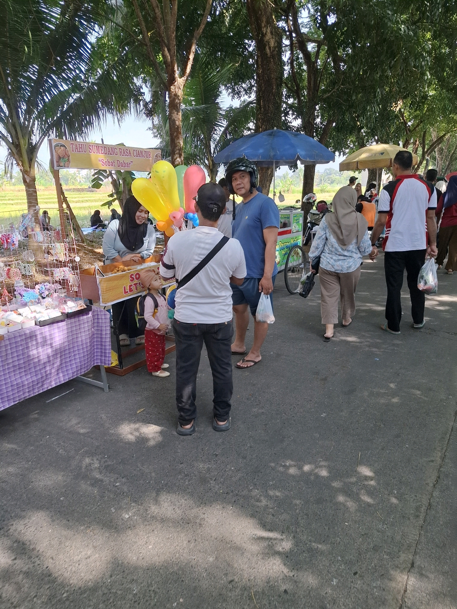 The image captures a street scene in Karangtengah, Indonesia, likely during an outdoor event or market. The foreground is dominated by a paved road shaded by trees, with dappled sunlight creating patterns on the asphalt.
On the left, a stall displays an assortment of colorful hair accessories. A woman in a grey hijab and a black top stands behind the stall, interacting with a young child wearing a pink long-sleeved shirt, red patterned pants, and a light brown hijab. The child carries a small backpack. Adjacent to this stall is another vendor's cart with a sign that reads "TAHU SUMEDANG RASA CIAMIS" and "Sobat Dahar". A bunch of colorful balloons, yellow, pink, and green, are attached to the cart.
In the mid-ground, a man wearing a blue t-shirt and a helmet is looking towards the camera. He is holding a white plastic bag. Next to him, a woman in a light blue patterned shirt and beige pants walks away from the camera, holding a black bag. Behind them, a man in a white t-shirt with red and black accents is walking, also carrying a white plastic bag. Further back, other individuals are visible, some walking and some standing, with a motorcycle and a bicycle parked nearby. A yellow umbrella shades another area.
The background reveals a rural landscape with rice paddies and lush green trees, suggesting a location on the outskirts of a more populated area. The overall atmosphere appears to be casual and lively, with people engaged in shopping and socializing under a bright sky.