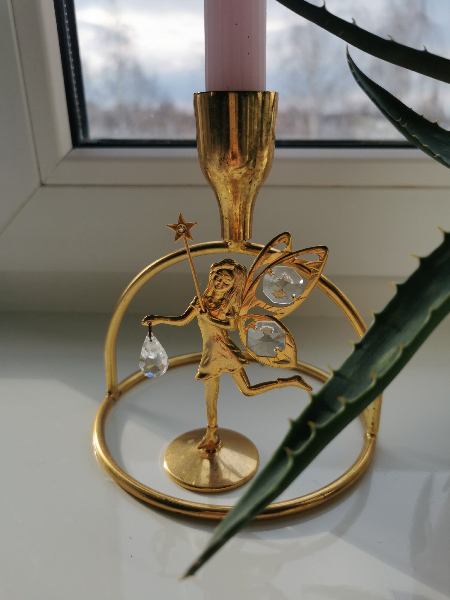 A close-up shot depicts a golden fairy figurine candlestick holder with a pink candle. The fairy figure has crystal wings and a star-topped wand, and is holding a crystal teardrop. The scene is set against a windowsill, with part of a green aloe vera plant on the right and a window with a view of a cloudy sky and bare trees in the background. The photograph has been taken indoors, likely in a home, on a white surface. The specific location within the city of Unknown, Unknown, is not discernible from the image.