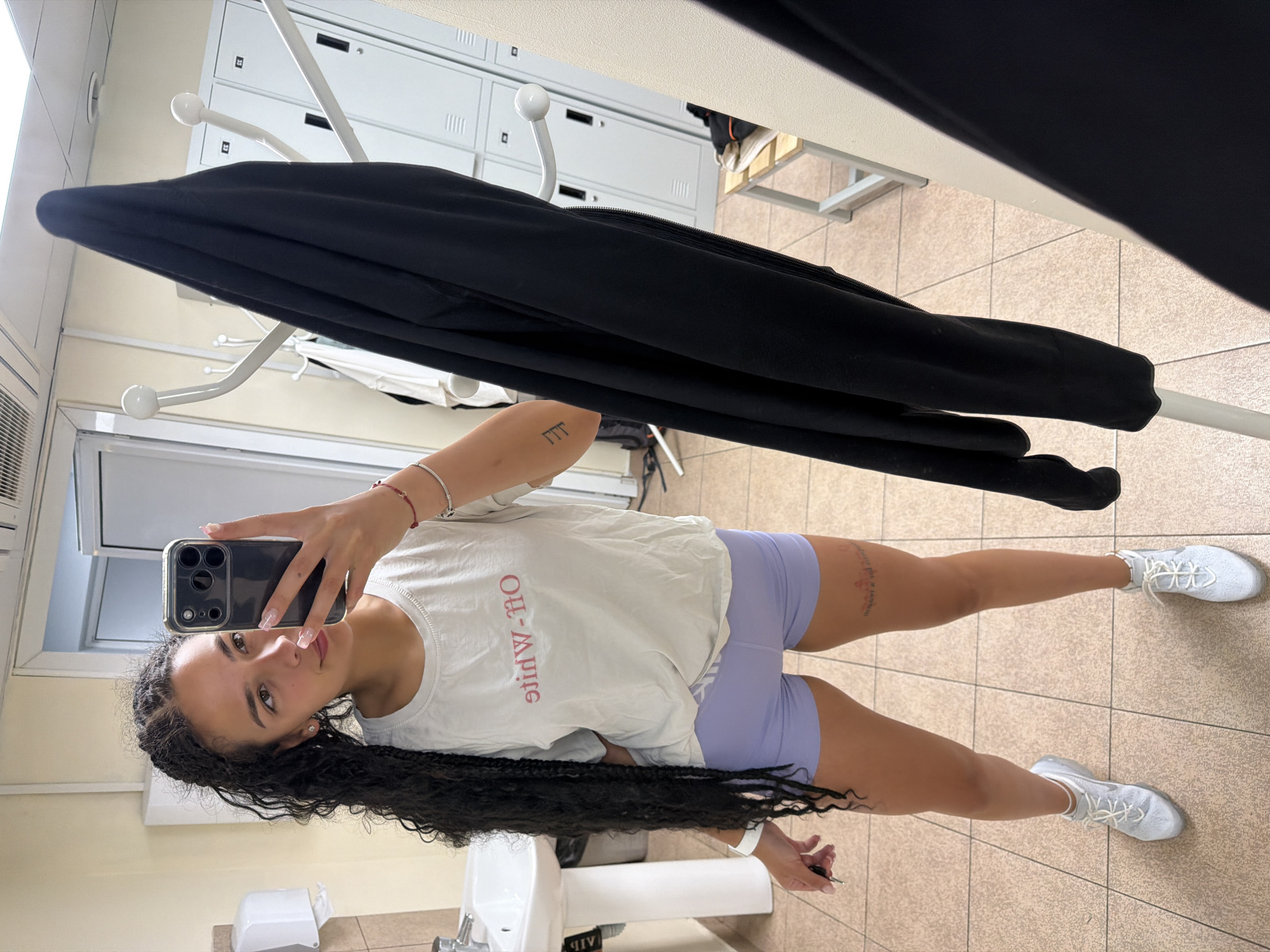 A young woman with long, dark, curly hair braided into a ponytail stands in what appears to be a locker room or changing area. She is taking a selfie with her phone, which is held in her right hand. She is wearing a light-colored t-shirt with "Off-White" printed on it in pink, and light purple athletic shorts. Her left hand is visible near her hip, holding a small item, possibly keys. She has a tattoo on her upper left thigh. Her footwear consists of light-colored athletic shoes. In the background, a black jacket hangs on a white coat rack. To the right of the coat rack, white lockers are visible. A sink and a paper towel dispenser are on the left side of the image. The floor is tiled with beige square tiles. The overall environment suggests a public facility, likely a gym or sports facility, but no specific location in Gabrovo, Bulgaria, can be determined from the visual information.