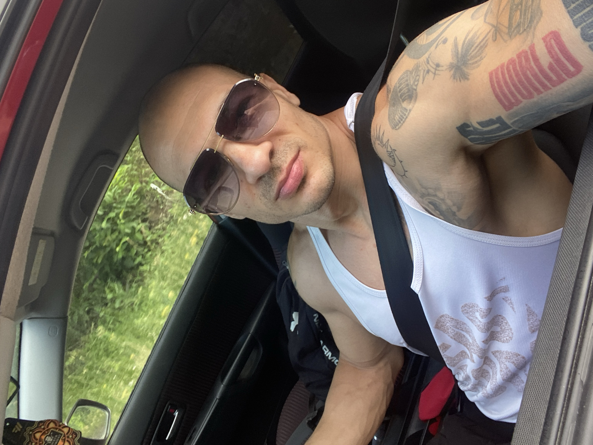 An adult male with a bald head and light skin tone is depicted in a selfie taken from the driver's seat of a vehicle, in or near Pleven, Bulgaria. The image does not contain specific visual landmarks to confirm this location. He is wearing dark aviator-style sunglasses with gold-toned frames and a white ribbed tank top with a light-colored, abstract pattern on the torso. A black seatbelt is securely fastened across his chest and lap. His right arm (viewer's left) is flexed, displaying prominent muscle definition and veins. Both arms are extensively tattooed; designs visible on his right arm include a large red rectangular pattern, dark vertical text, palm trees, a microphone, and a seashell.
The car's interior is predominantly dark, featuring a black door panel with a silver-colored handle and a red accent light on the driver's side. An ornate, orange-patterned air freshener hangs from the rearview mirror. A dark garment, possibly a hoodie, is crumpled on the passenger seat or behind the driver, and a partial view of a red object (likely fabric or a bag) is visible on the seat below his left arm. A strip of red trim is visible along the top interior edge of the vehicle's frame.
The background, viewed through the driver's side window and windshield, consists of highly blurred green foliage, indicative of motion through a natural or rural roadside environment during daylight hours.