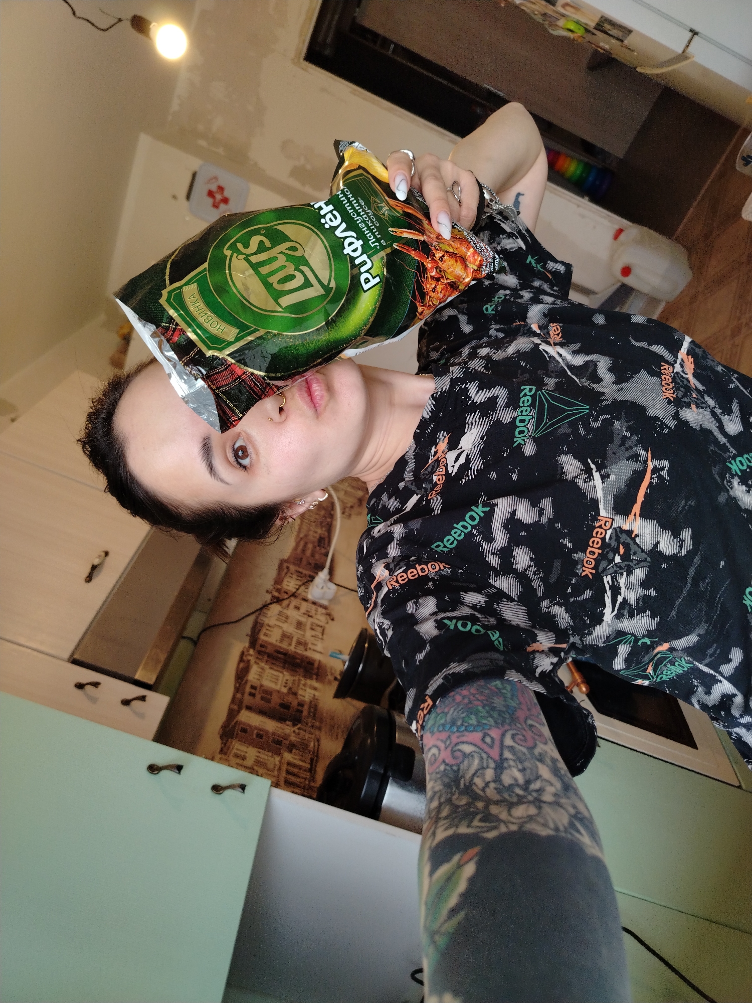 A woman is holding a bag of Lays potato chips in front of her face in what appears to be a kitchen setting. The bag of chips is green and has the text "Рифлён", "Лангустин в пикантном coyce", and "НОВИНКА". The woman is wearing a black Reebok shirt with white and orange patterns. Her left arm is heavily tattooed. The kitchen has light green cabinets with bronze-colored handles, and the backsplash features a cityscape design. A lightbulb hangs from the ceiling. There's a white object near a set of rainbow-colored cylinders. The location is identified by the Cyrillic text on the chip bag.