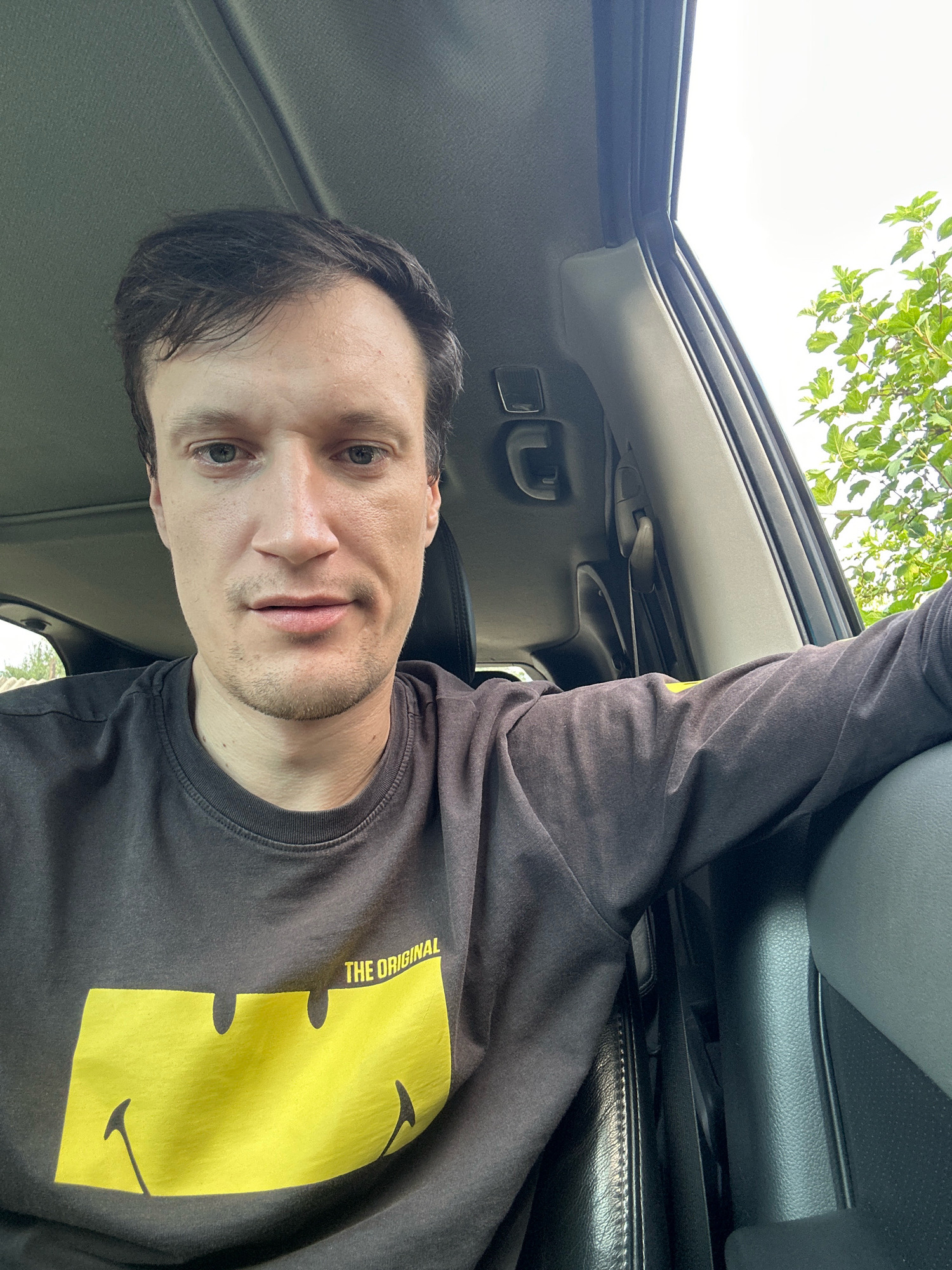 A man is sitting in the driver's seat of a vehicle, looking at the camera. He is wearing a dark gray long-sleeved t-shirt with a large yellow smiley face graphic on the front, and the words "THE ORIGINAL" printed above it. His left arm is resting on the car's interior. The interior of the car appears to be black and gray. Outside the car window, green foliage is visible against a bright sky. The specific location in Shatava, Ukraine is not discernible from the image.