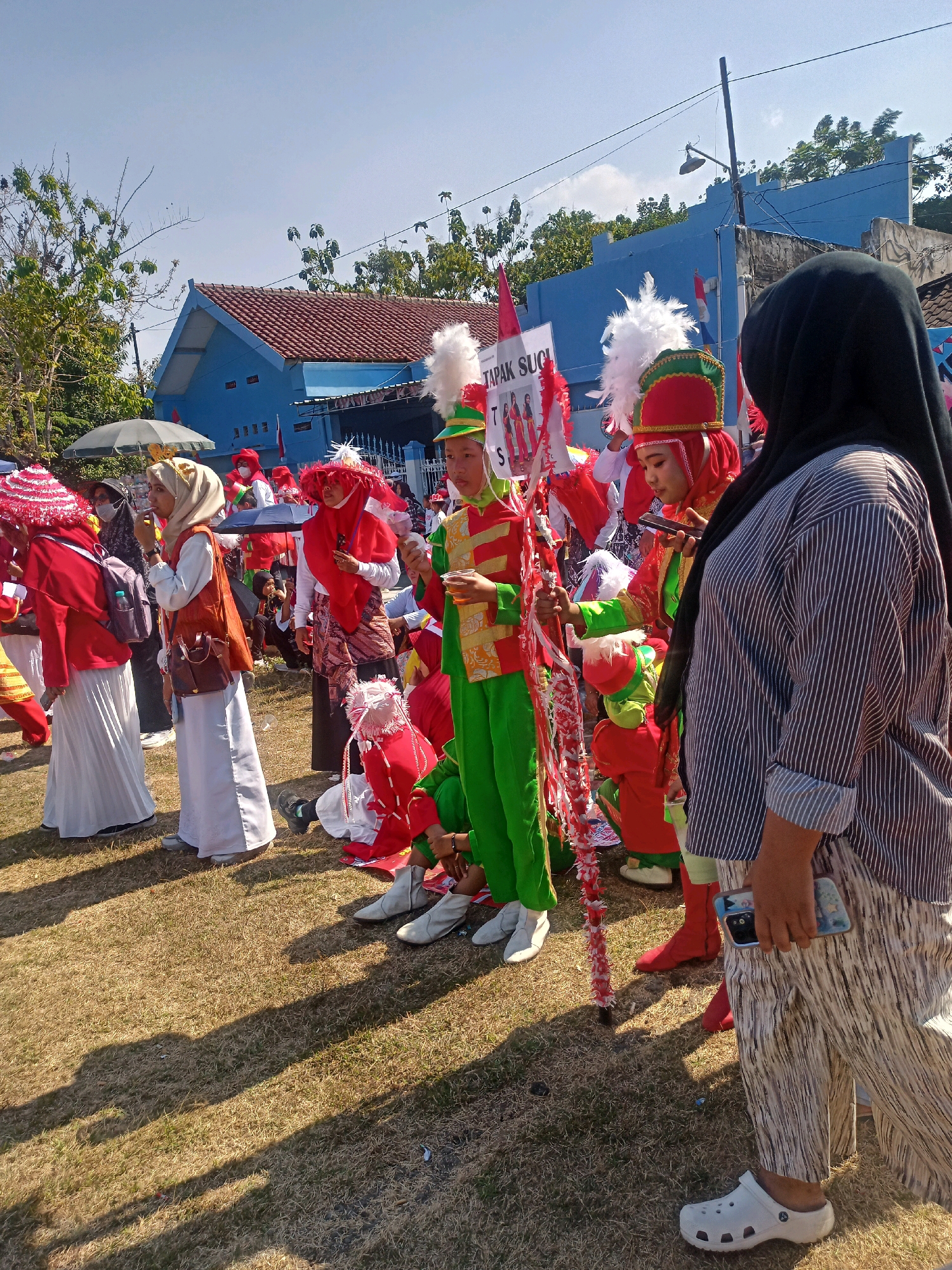 In Sumberlawang, Indonesia, on a sunlit day, a group of people, predominantly young individuals in vibrant marching band attire, are gathered on a dry, grassy field. The performers are dressed in red and green uniforms with elaborate hats adorned with white plumes. Some carry banners and decorative poles, suggesting a parade or performance. One individual holds a sign with text that appears to be in Indonesian. In the foreground, a woman wearing a black hijab and a striped shirt stands to the right, looking at her phone. In the background, a blue building with a red-tiled roof and trees can be seen. The overall atmosphere appears festive and community-oriented.