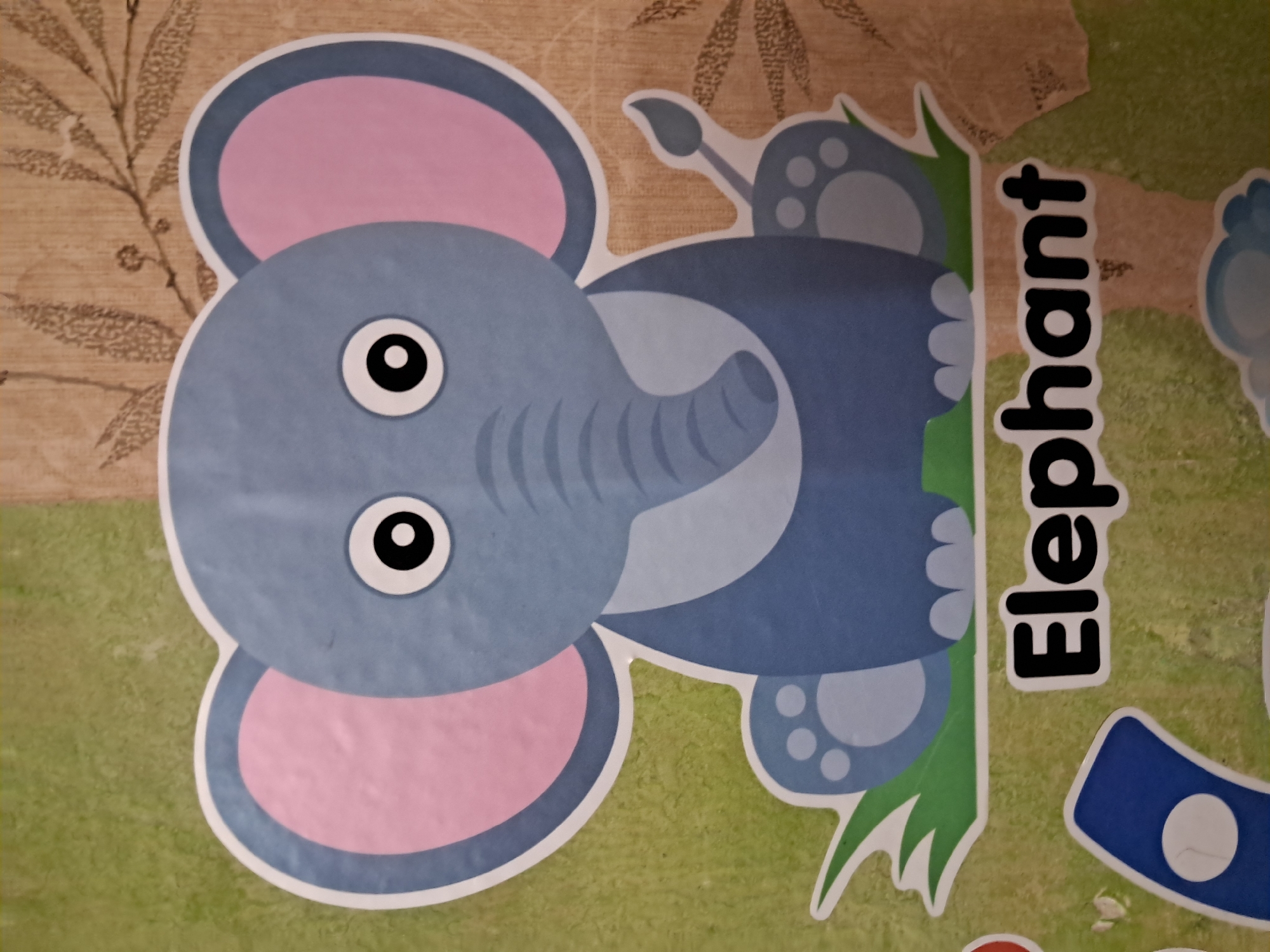 The image shows a cartoon illustration of an elephant. The elephant is blue with large pink ears and black eyes. It is sitting on green grass, and the background is a textured brown material with a faint pattern of leaves. Below the elephant, the word "Elephant" is written in large black letters with a white outline. The image appears to be a decal or sticker, possibly for educational purposes. The location is unknown, Unknown.