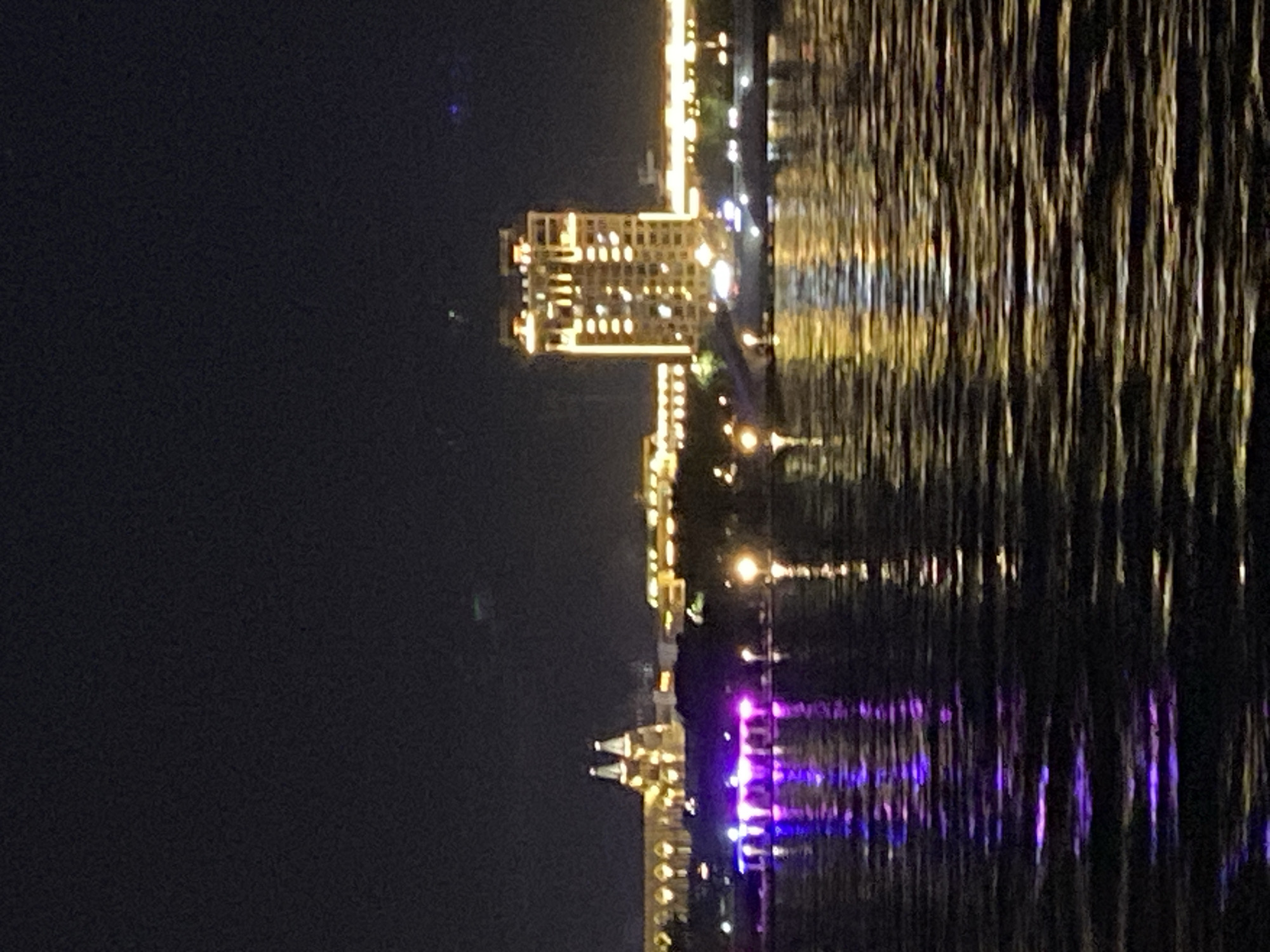 The image depicts a nighttime scene of the Blagoveshchensk city embankment along the Amur River. In the distance, illuminated buildings and structures line the riverfront. A prominent, multi-story building with numerous lit windows stands out, appearing to be a hotel or office building. To the left of this building, there is a structure with arched windows and two conical towers, possibly a historical or cultural building. The embankment area is lit with colorful lights, particularly purple lights reflecting on the water in the foreground. The water surface shows ripples and reflections of the city lights. The sky is dark, indicating nighttime. There are no people or vehicles clearly visible in the image. The overall scene conveys a view of a well-lit urban waterfront at night.