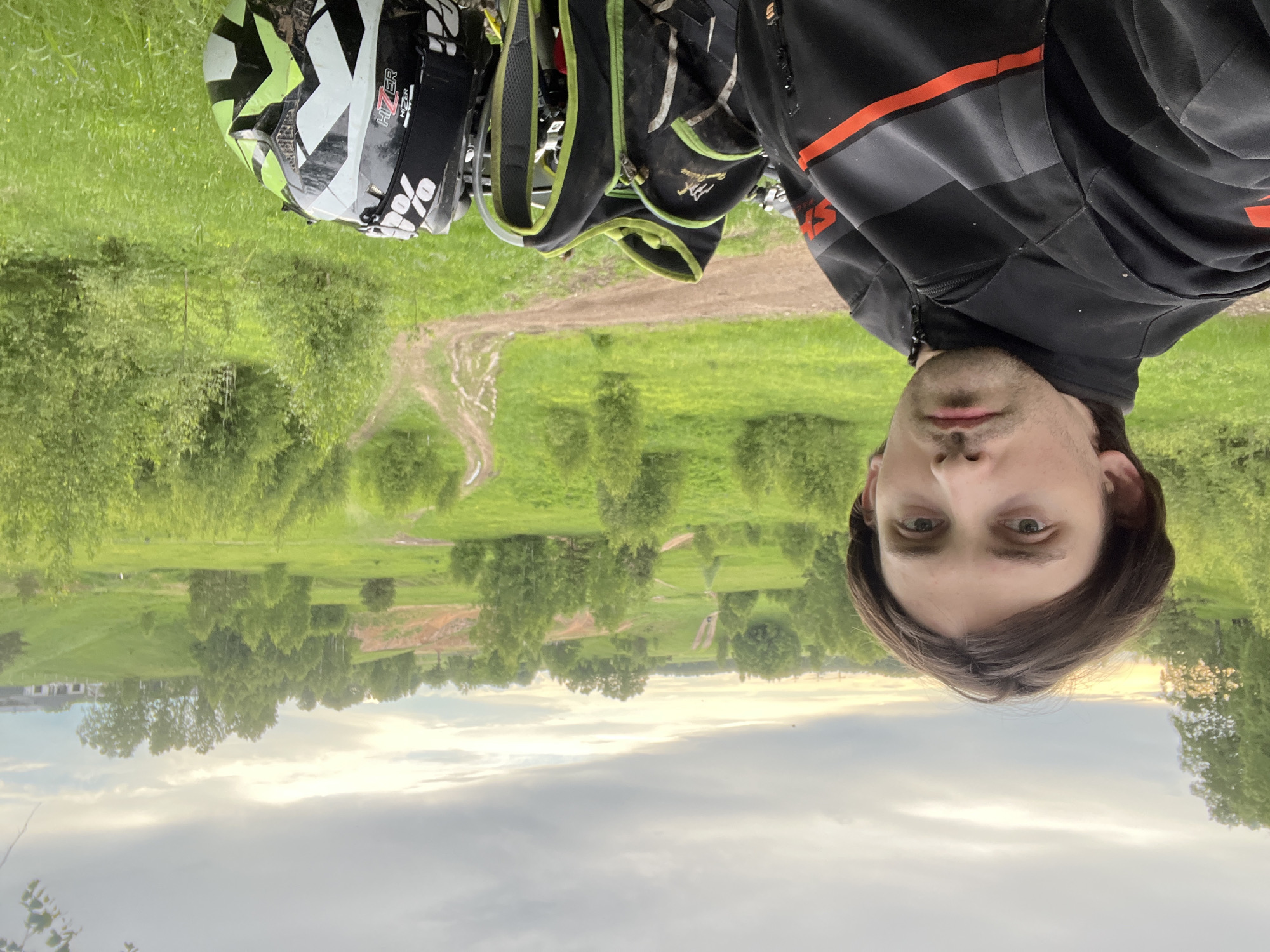 A man stands in Новочувашский, Russia. He has brown hair and is wearing a black jacket with orange stripes. Behind him is a grassy field with trees and a dirt path. A helmet and backpack are also visible in the image.
