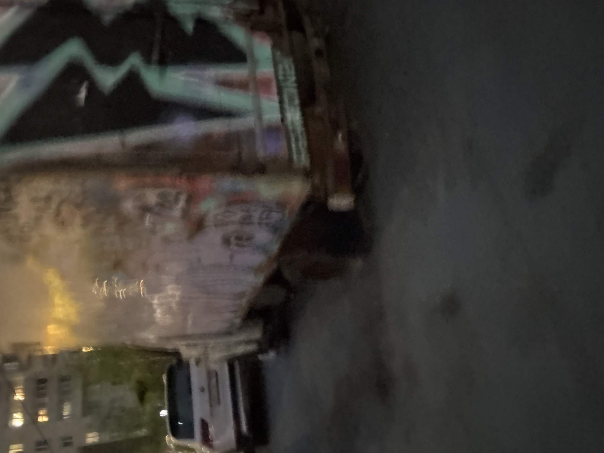 The image depicts a street scene in Saint Petersburg, Russia, likely at night or in low light. A heavily graffitied truck or large vehicle occupies the left side of the frame, with its front end extending into the foreground and its rear partially visible in the background. A white car is parked behind the truck. In the distance, a multi-story building is visible, with lit windows suggesting residential or commercial occupancy. The street appears paved, with some darker patches that could be shadows or stains. The overall environment suggests an urban setting, possibly an industrial or less affluent area, judging by the graffiti and the general state of the vehicle. There are no discernible people or active interactions captured in the image.