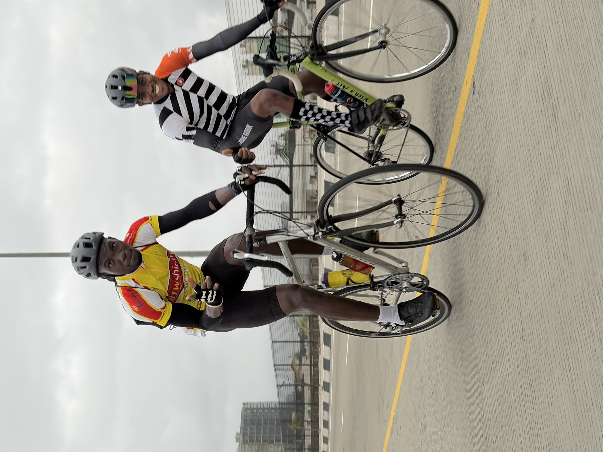 Two cyclists, both wearing helmets and cycling gear, ride on a paved road. The cyclist in the foreground on the left wears a yellow and red jersey and black shorts, with black gloves. The cyclist on the right wears a black and white striped jersey with orange accents and black shorts, also with black gloves. Both cyclists are on road bikes. The road has a yellow line marking on the right side. In the background, there is a fence, some buildings under construction, and a cloudy sky. The scene appears to be in Alaguntan, Nigeria, a coastal region known for its urban development and proximity to water, which could be inferred from the presence of construction and the elevated road structure suggesting infrastructure development in the area.