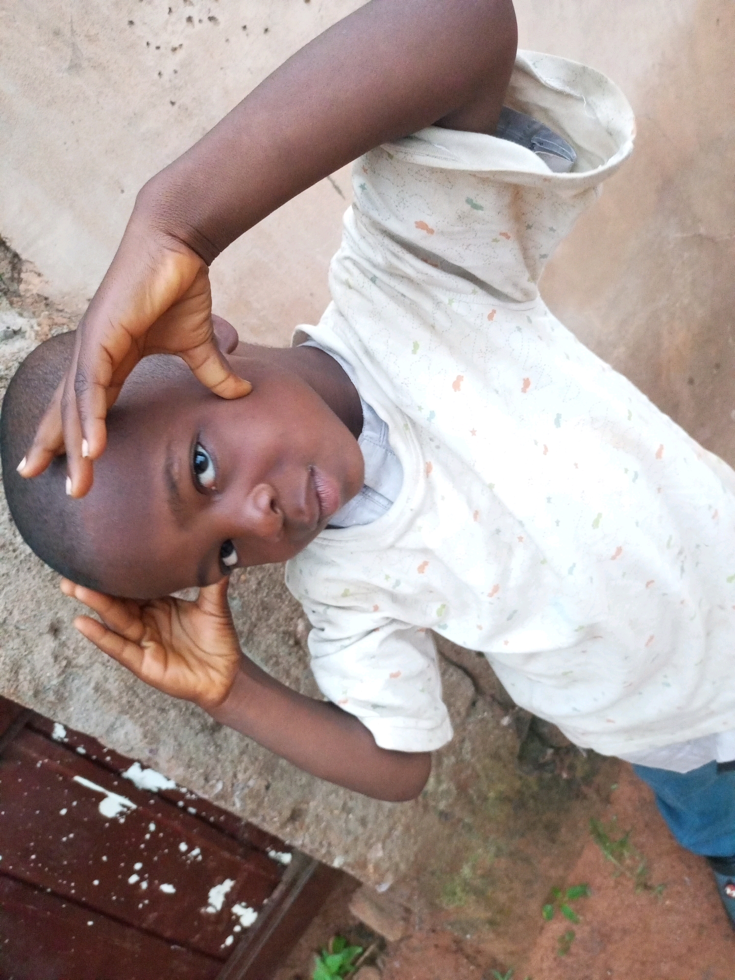 A young Black child is lying on their back outdoors, with their head tilted back and hands held up to their face. They are wearing a white t-shirt with a subtle pattern of small orange, green, and blue shapes. The child's skin is dark brown, and their eyes are open, looking directly at the camera. The background appears to be dirt ground with some scattered pebbles and a hint of brown wooden material to the left. The overall lighting suggests it is daytime. The scene is likely in a residential area of Tempure, Nigeria.