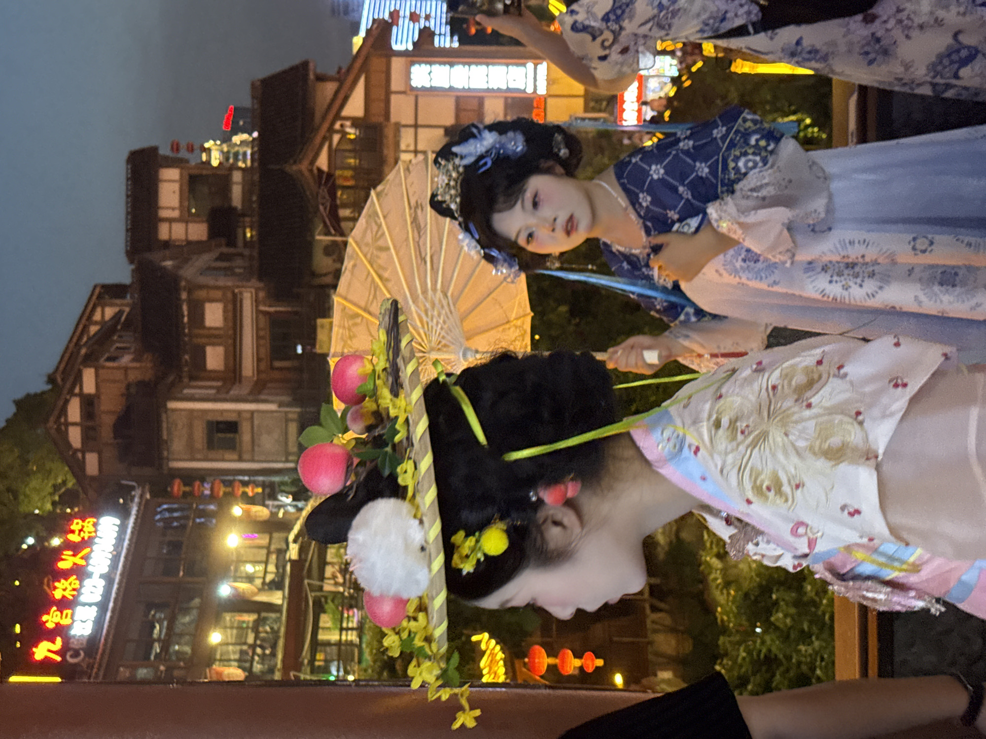 In the Yuzhong District of China, at night, two young women dressed in traditional Chinese attire stand side-by-side. The woman on the left wears a floral patterned robe and a large, ornate headpiece adorned with faux fruit and flowers. She is looking to the left, her profile visible. The woman on the right is holding a closed, tan umbrella, her face slightly turned towards the viewer. She wears a blue and white patterned dress with puffy sleeves and a pearl necklace. In the background, a bustling cityscape is visible with illuminated buildings and traditional Chinese architecture. The scene is lit by the glow of streetlights and neon signs.