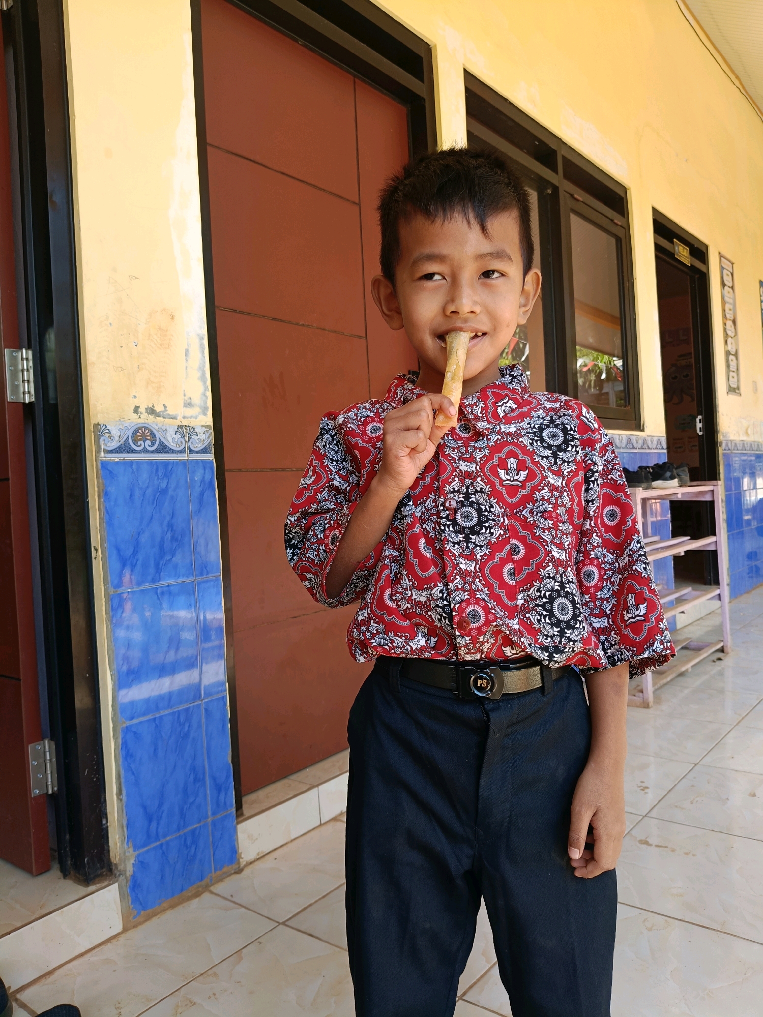 An Indonesian boy with short black hair and light brown skin is standing outside and holding a stick of food to his mouth. He is wearing a red and white patterned shirt and dark pants with a black belt that has the letters "PS" on it. The boy is looking to his left with a slight smile. He is standing in front of a building with yellow walls and brown doors. There are blue tiles on the wall next to the door. The ground is tiled with light brown and white squares. The scene appears to be in a rural or suburban area.