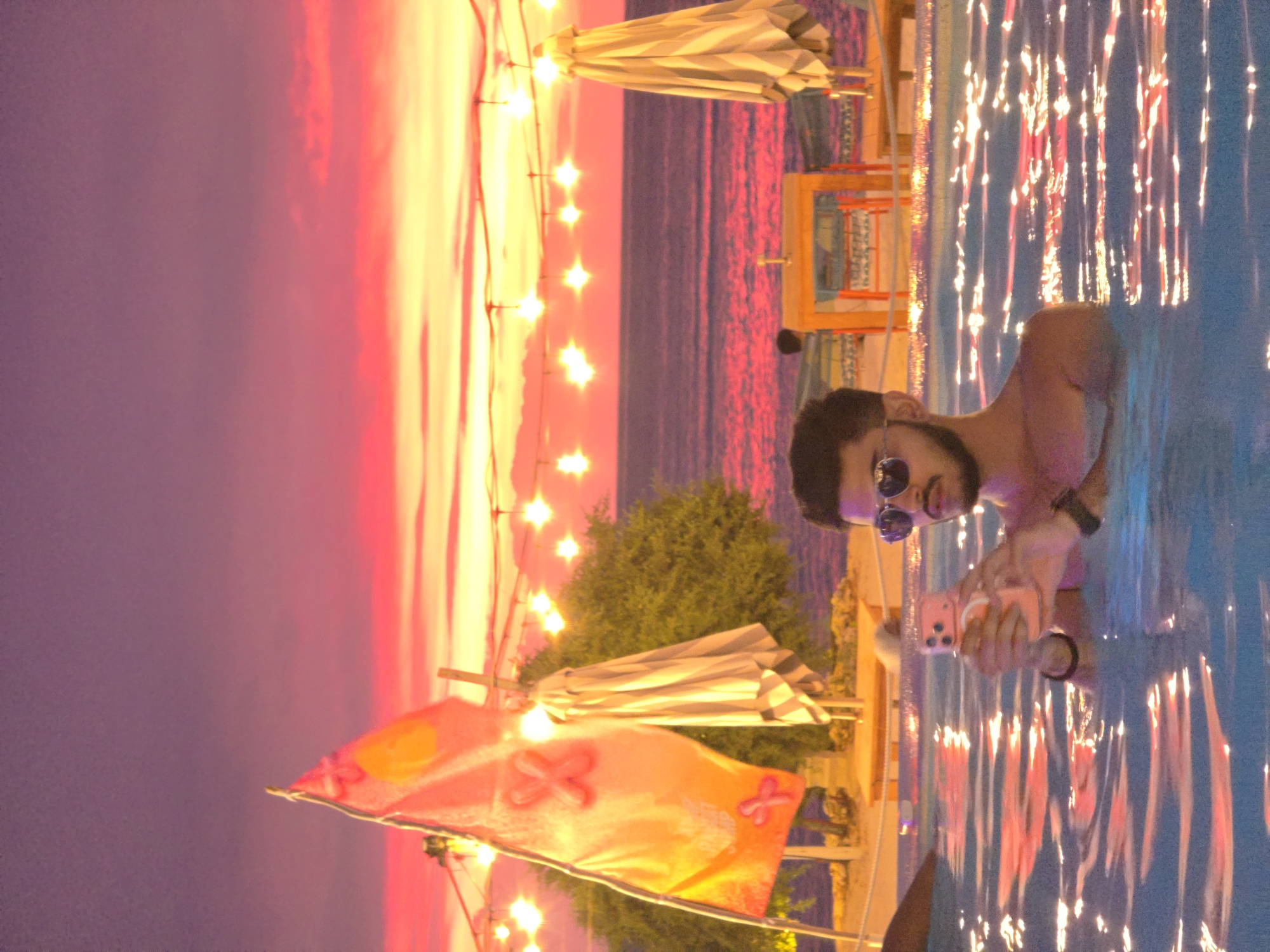 A medium indoor shot shows a man waist-deep in a swimming pool, illuminated by sparkling lights on the pool's surface, taken in Unknown, Unknown. The man, who has dark hair, a beard, and is wearing sunglasses, is looking at the camera while holding a pink smartphone with his right hand. A wristwatch is visible on his left wrist. In the background, a body of water, possibly the ocean or a large lake, reflects a vivid pink sky at sunset. Strings of small, star-shaped lights hang vertically, adding to the ambiance. There is a beach umbrella, various chairs, and a decorative orange flag. The general scene suggests an outdoor setting near a beach or resort.