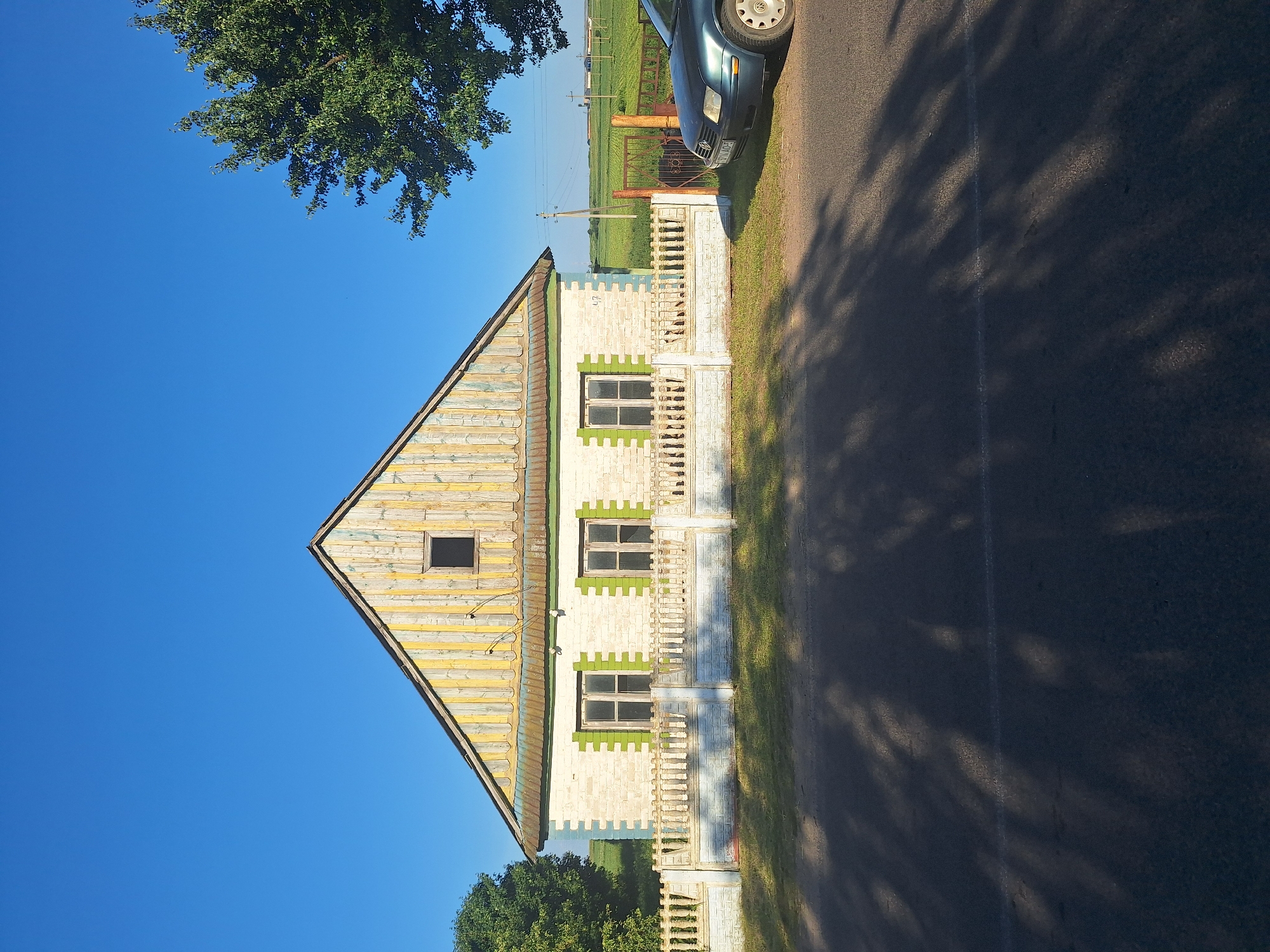 The image shows a single-story house with a gabled roof, located in Цімкавіцкі сельскі Савет, Belarus. The house has a white brick exterior with green accents around the windows. The roof is made of weathered, vertical wooden planks painted in alternating yellow and white stripes. A small, dark square window is visible in the attic. A low, decorative fence made of white brick and posts surrounds the front of the property. Two windows with green shutters are visible on the front of the house. A dark-colored car is parked on the right side of the image, partially obscuring a metal gate and fence leading to a green field. Trees are present on the left and right sides of the house, and utility poles are visible in the background. The sky is clear and blue, and shadows from trees are cast across the asphalt road in the foreground.