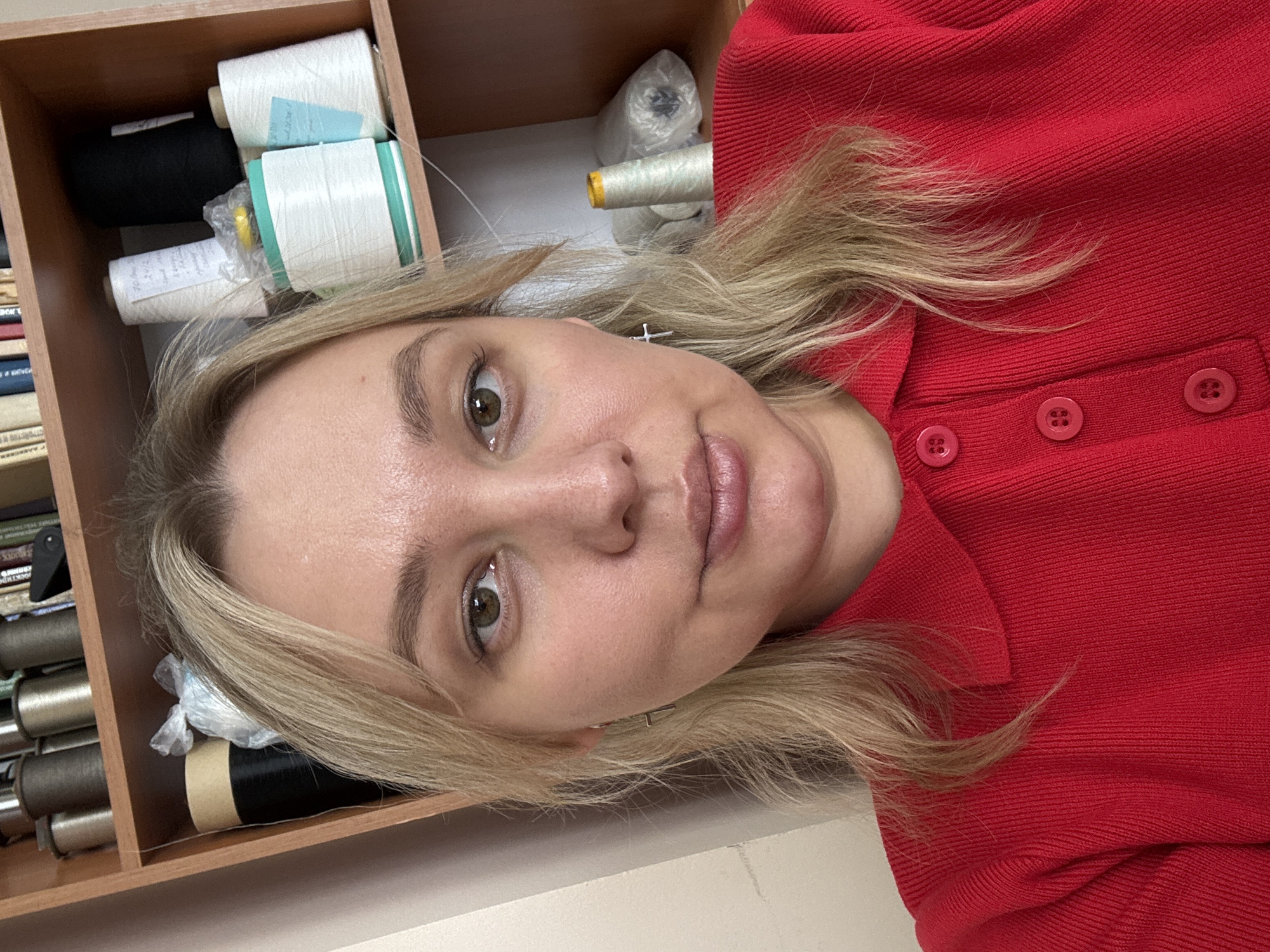 A blonde-haired Caucasian woman, appearing in a direct frontal selfie, is centered in the frame. She has light skin, greenish-brown eyes, and is wearing a red, collared, ribbed knit shirt with three visible red buttons down the placket. A silver-toned cross earring is visible on her right ear. Her hair is parted slightly to the left and falls to her shoulders. Behind her is a light brown wooden shelving unit. The top shelf holds several spools of thread in varying shades of brown and gold, alongside some books. The middle shelf contains large spools of black and white thread, one with a teal base, another with a yellow top, and various smaller spools, some bearing handwritten labels. Clear plastic bags containing white materials are also present. The lower right shelf shows more spools of light-colored thread and a rolled item in a plastic bag. This scene is located in Chaikovsky, Russia.