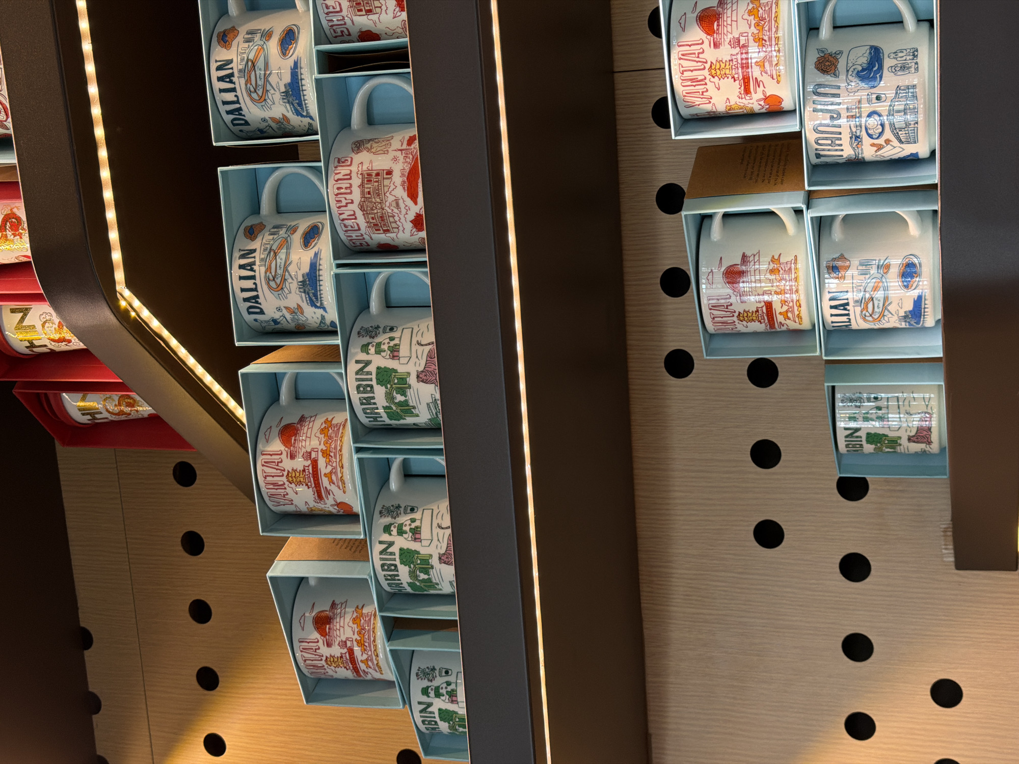 The image shows a display of city-themed mugs in individual light blue boxes. The mugs feature designs and names of various cities including "DALIAN," "SHENYANG," "HARBIN," "YANTAL," "TIANJIN" and "UTAL". They are arranged in a stacked configuration within a dark gray display unit highlighted with vertical LED strips. The background is a light wood panel with evenly spaced, large black circles.
