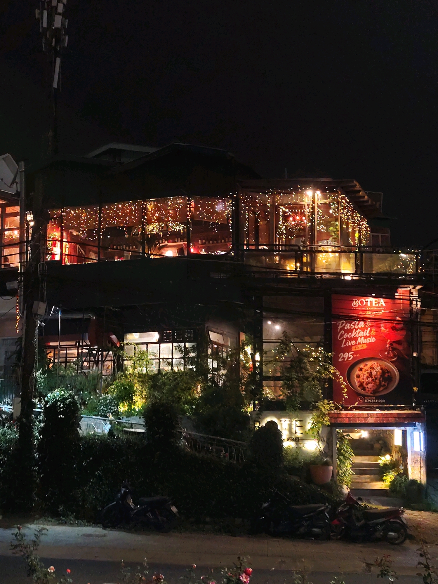 The image captures a multi-story building at night in Da Lat, Vietnam. The building, likely a restaurant or cafe named "BOTEA," is adorned with numerous string lights on its exterior, creating a warm and inviting ambiance. A large red banner on the right side of the building advertises "Pasta, Cocktail & Live Music" with a price of "295++." The lower levels of the building feature large glass windows, revealing interior lighting and glimpses of seating areas. Lush green foliage and bushes surround the ground level, with a few motorcycles parked in front. The sky is dark, indicating nighttime. The overall scene suggests a lively evening spot in Da Lat.