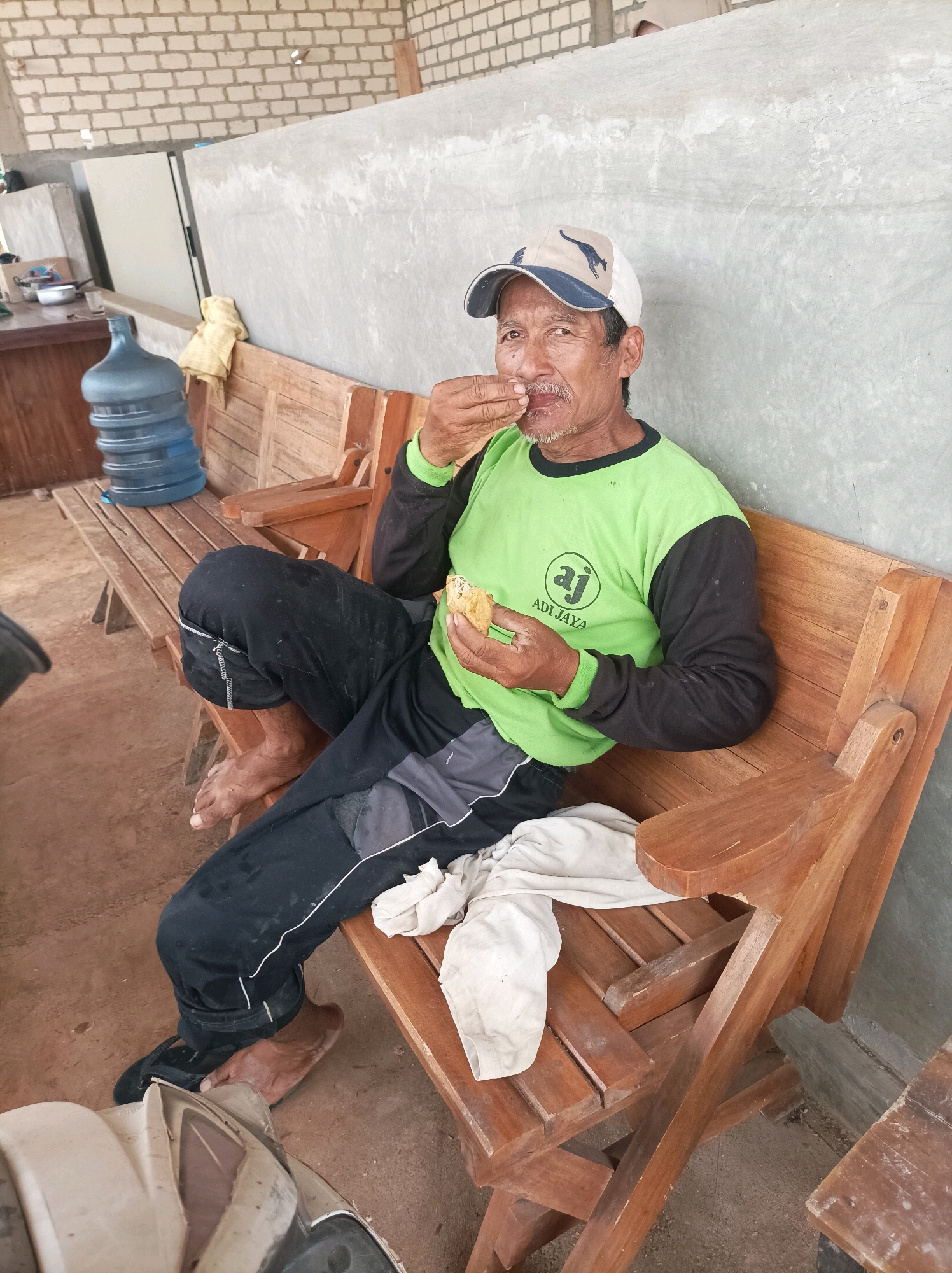 A man in a green and black long-sleeved shirt and black pants is sitting on a wooden bench, eating. He is wearing a baseball cap. In the background, there is a large blue water jug and a wall made of concrete blocks. The scene appears to be outdoors, possibly in Merakurak, Indonesia. The man is holding a piece of food to his mouth and is looking towards the camera. The bench is made of wooden slats and has a worn appearance. A white cloth is draped over the bench.