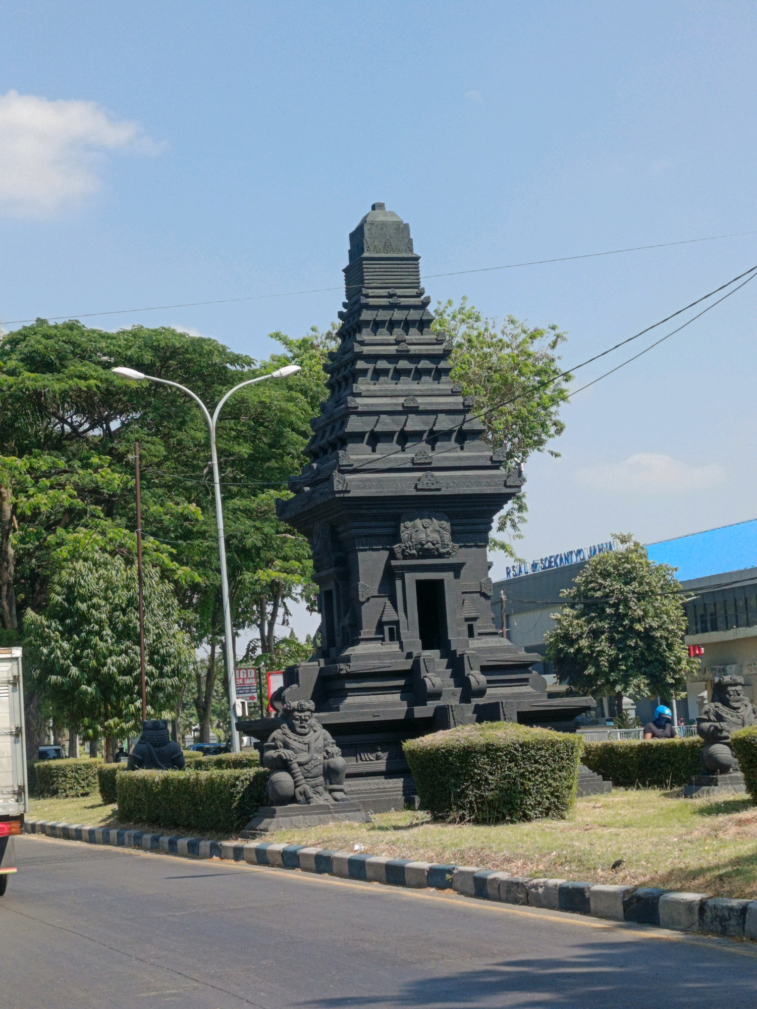 A dark stone temple structure stands tall on the right side of the image. It is intricately carved with multiple tiers and features a small opening at its base. In front of the temple, on either side of a manicured hedge, are two stone statues of seated figures. A modern street light with two lamps is visible on the left, positioned in front of lush green trees. A paved road runs across the foreground, with a truck partially visible on the far left. In the background, a building with a bright blue roof is seen, bearing the text "RSAL dr. SOEKANTYO JAH". The sky above is clear blue with a few wispy clouds. The scene appears to be located in an urban area in Sedati Agung, Indonesia.