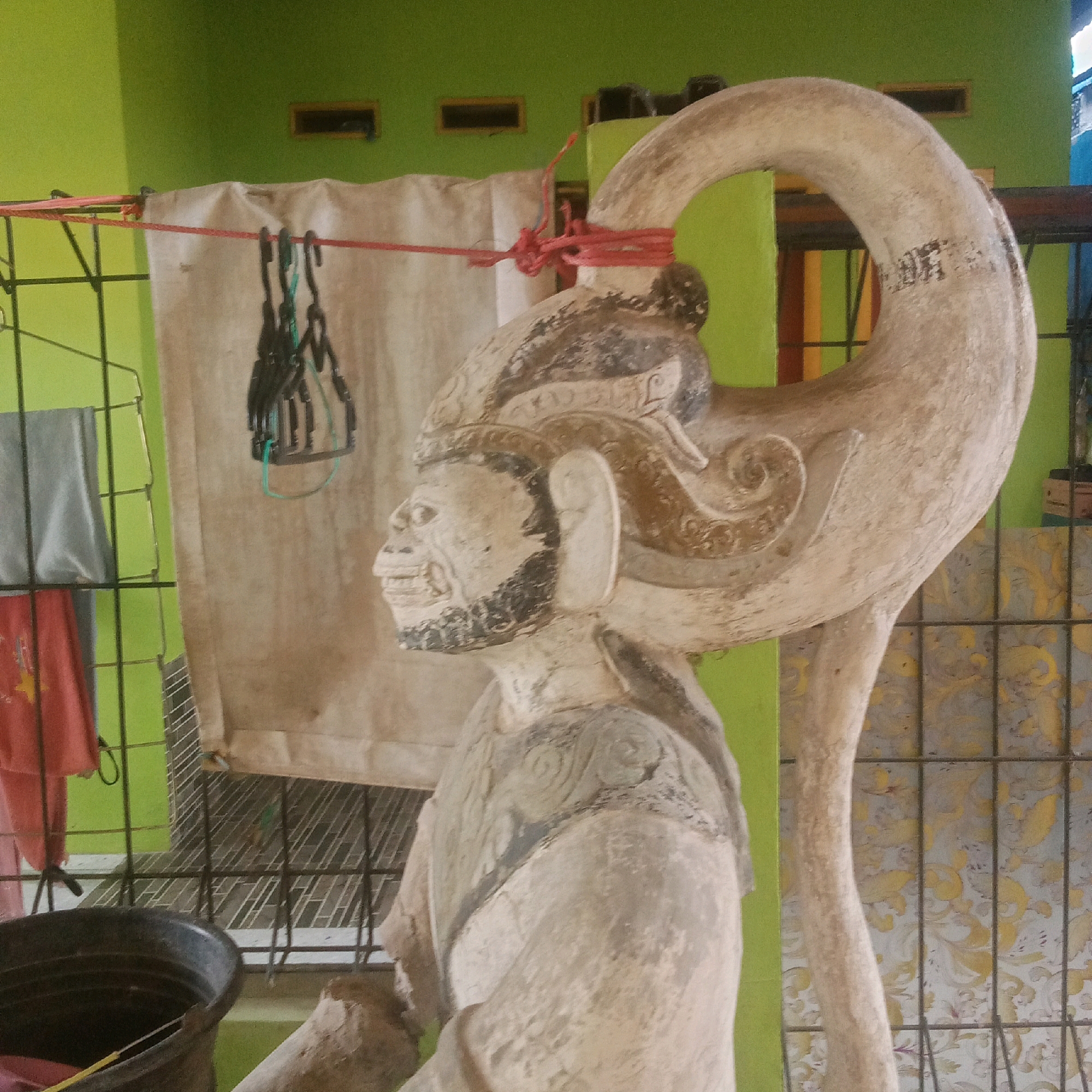 An outdoor scene in Karawang, Indonesia features a light-colored, weathered statue of a mythical creature with a human-like face and a long, curved horn or tail. The statue is partially wrapped with red twine and is positioned in front of a green wall and a patterned fabric backdrop. To the left of the statue, a black plastic bucket and some folded fabric are visible. A metal rack with hanging clothes is also present. The ground appears to be covered with gravel and dirt, and the overall setting suggests a utilitarian or storage area.