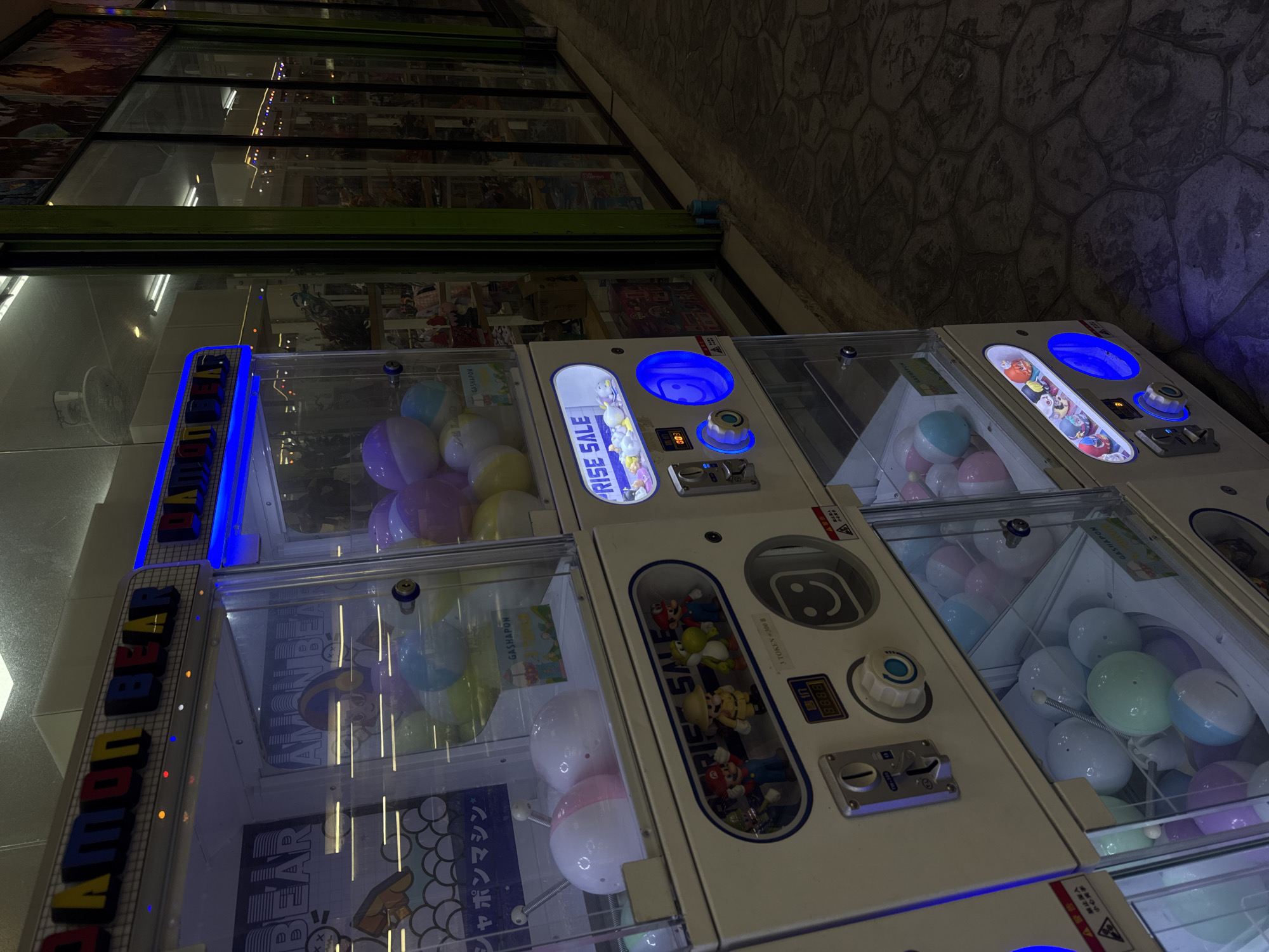 This media file shows a row of gumball or capsule toy vending machines, likely located in a brightly lit entertainment area or arcade at night. The machines are filled with various colorful capsules and some appear to contain small prizes like figurines. The vending machines have prominent labels, some of which are in Japanese, reading "GASHAPON" and "SHAPON MACHINE." One machine has a "RISE SALE" sign with small toys visible inside, and another shows "BEAR" in large blue lettering. The machines are illuminated by blue and other colored lights, creating a vibrant and inviting atmosphere. A textured stone wall is visible in the background, suggesting an outdoor or semi-outdoor setting, possibly a street or walkway in Karon, Thailand. The overall scene conveys a sense of fun and entertainment, typical of a vacation or entertainment district. There are no people visible in the frame.