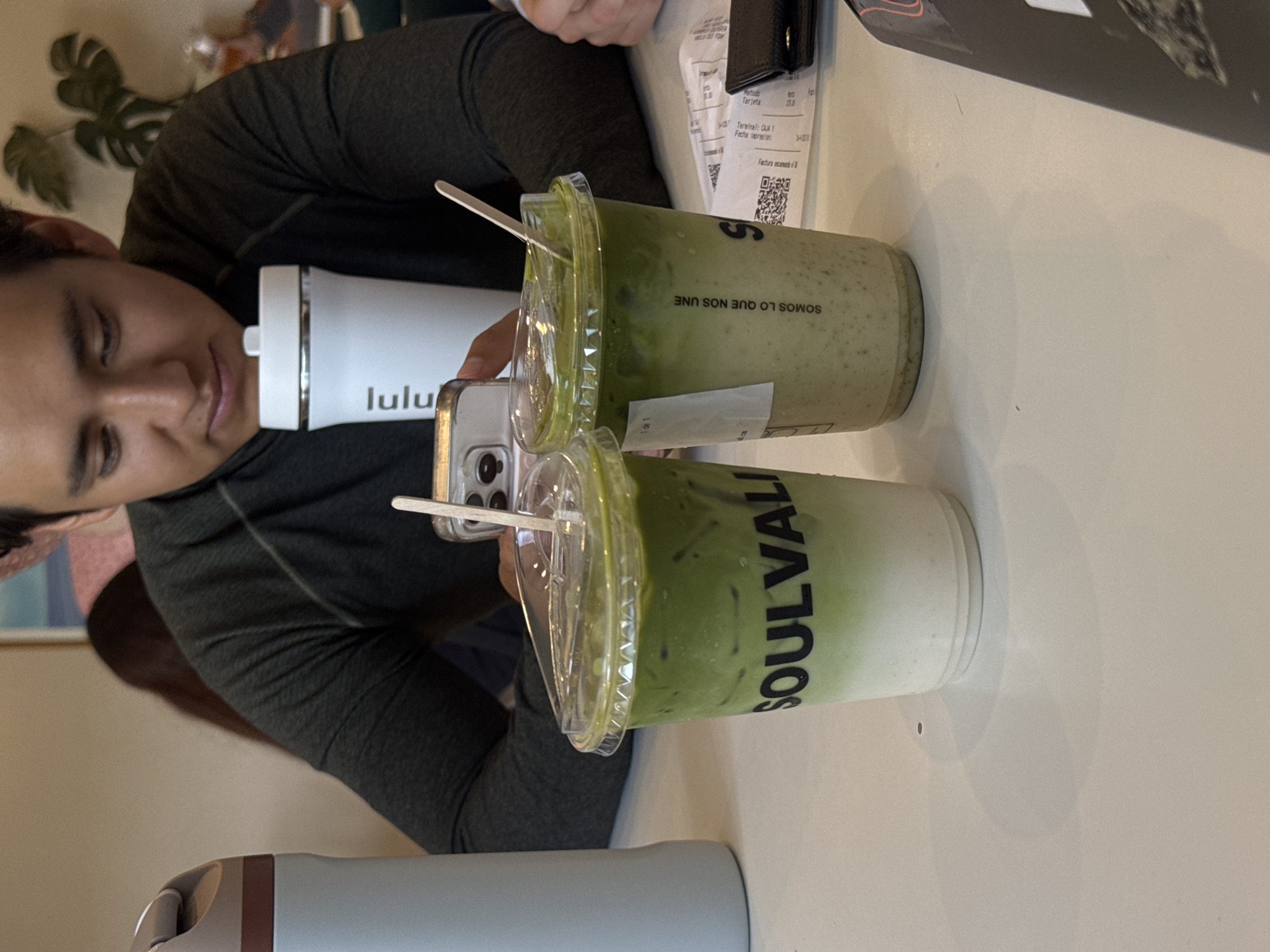 The image, captured in Puebla, Mexico, shows a person seated at a light-colored table, likely in a cafe or casual dining setting. The individual, appearing to be male, is seen from the chest up, with their head slightly tilted and a relaxed expression on their face. They are wearing a dark long-sleeved athletic-style top.
On the table in front of them are two clear plastic cups filled with a layered green beverage, likely a matcha latte, topped with ice and fitted with clear plastic lids. Each cup contains a white stirrer. One cup clearly displays the brand name "SOULVALI" vertically in black letters. The other cup, closer to the person, features the Spanish phrase "SOMOS LO QUE NOS UNE" (We are what unites us) printed upside down. A white water bottle with the "lulu" (Lululemon) logo is positioned next to the drinks.
The person is holding a smartphone, which is partially visible between the water bottle and one of the beverage cups. To the right of the drinks, a receipt is visible, displaying text such as "Metodo Tarjeta" (Card Method), "Terminal: CUA 1", "Fecha lxpresiat:", and "Factura escaneri" (Scanned invoice). A black wallet or small item is also on the table near the receipt. In the background, out of focus, a green plant and hints of indoor decor are discernible. The scene is illuminated by ambient indoor lighting.