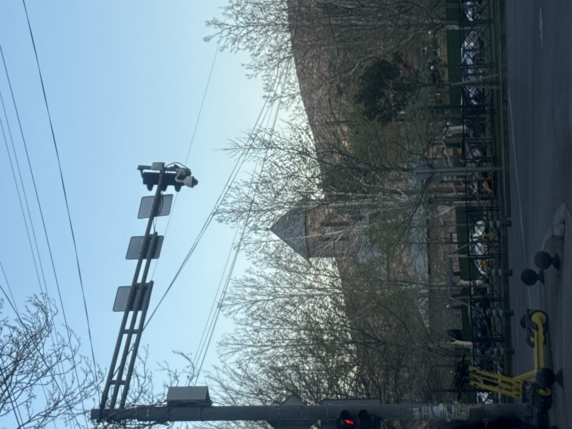 The image depicts an urban street scene in Tbilisi, Georgia, likely during late winter or early spring given the bare tree branches. The perspective is looking across a street, possibly from a vehicle.
In the foreground, a large utility pole stands prominently, supporting multiple overhead wires that crisscross the clear, light blue sky. Attached to this pole are a series of square traffic light signals, with the lowest visible light displaying red. What appear to be surveillance cameras are also mounted on the pole structure.
The midground features a line of trees with thin, leafless branches. Peeking through and above these trees, a building with a textured, possibly stone or brick, facade and a sloped, peaked roof is partially visible.
To the right of the scene, a paved street or sidewalk runs horizontally. Adjacent to this, a black metal railing or fence separates the paved area from what seems to be a grassy strip or wider pavement. Parked along this railing are several vehicles, including distinct bright yellow electric scooters or bicycles in a rack.
The overall scene suggests a quiet daytime moment with clear weather. No individuals are visible. There is no legible text present on any signs or objects within the frame.