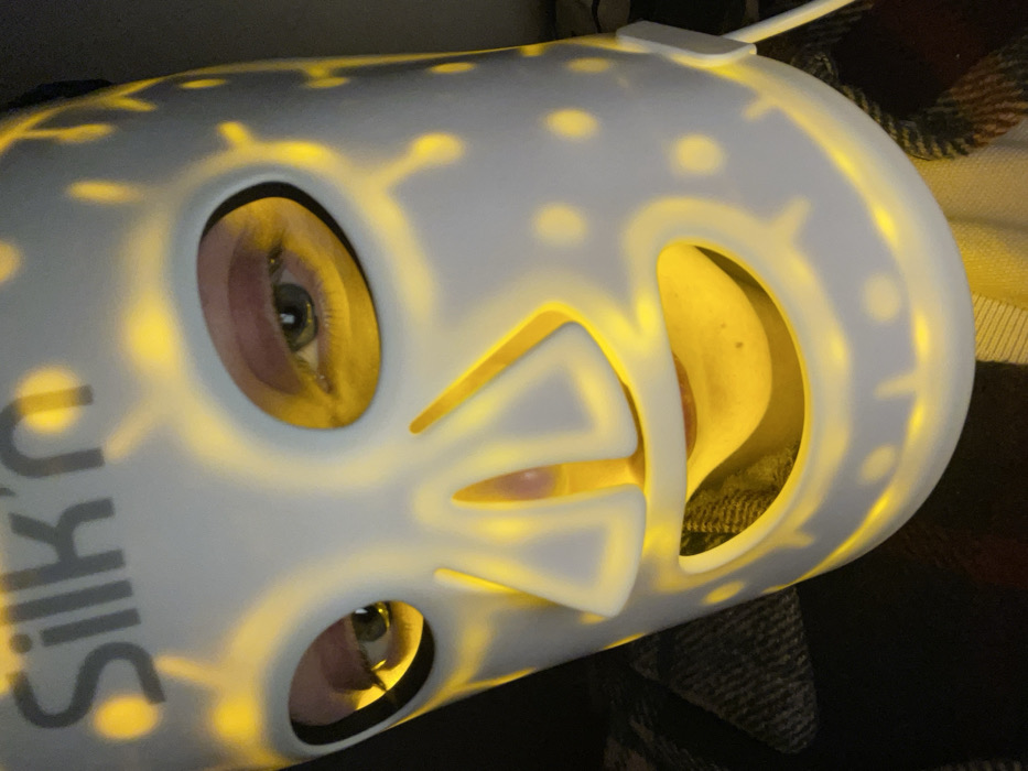 A person is depicted in a close-up, angled view, wearing a white LED light therapy mask. The mask, bearing the grey "Silk'n" brand logo across the forehead, emits a warm yellow light that illuminates the wearer's face through oval eye cutouts, a stylized nose aperture, and a broad smile-like mouth opening. Through these openings, the person's light green/hazel eyes are visible, looking forward. The bridge and tip of the nose, as well as the chin and mouth area, are also discernible beneath the mask's glow. Small circular perforations and patterned lines on the mask's surface transmit the yellow light externally. A white power cable extends from the right side of the mask. The immediate background is dark, with a textured, dark-patterned fabric, possibly a blanket or clothing, visible below and to the left of the mask. The specific location within Monsummano Terme, Italy, cannot be determined from the visual information provided in the image.