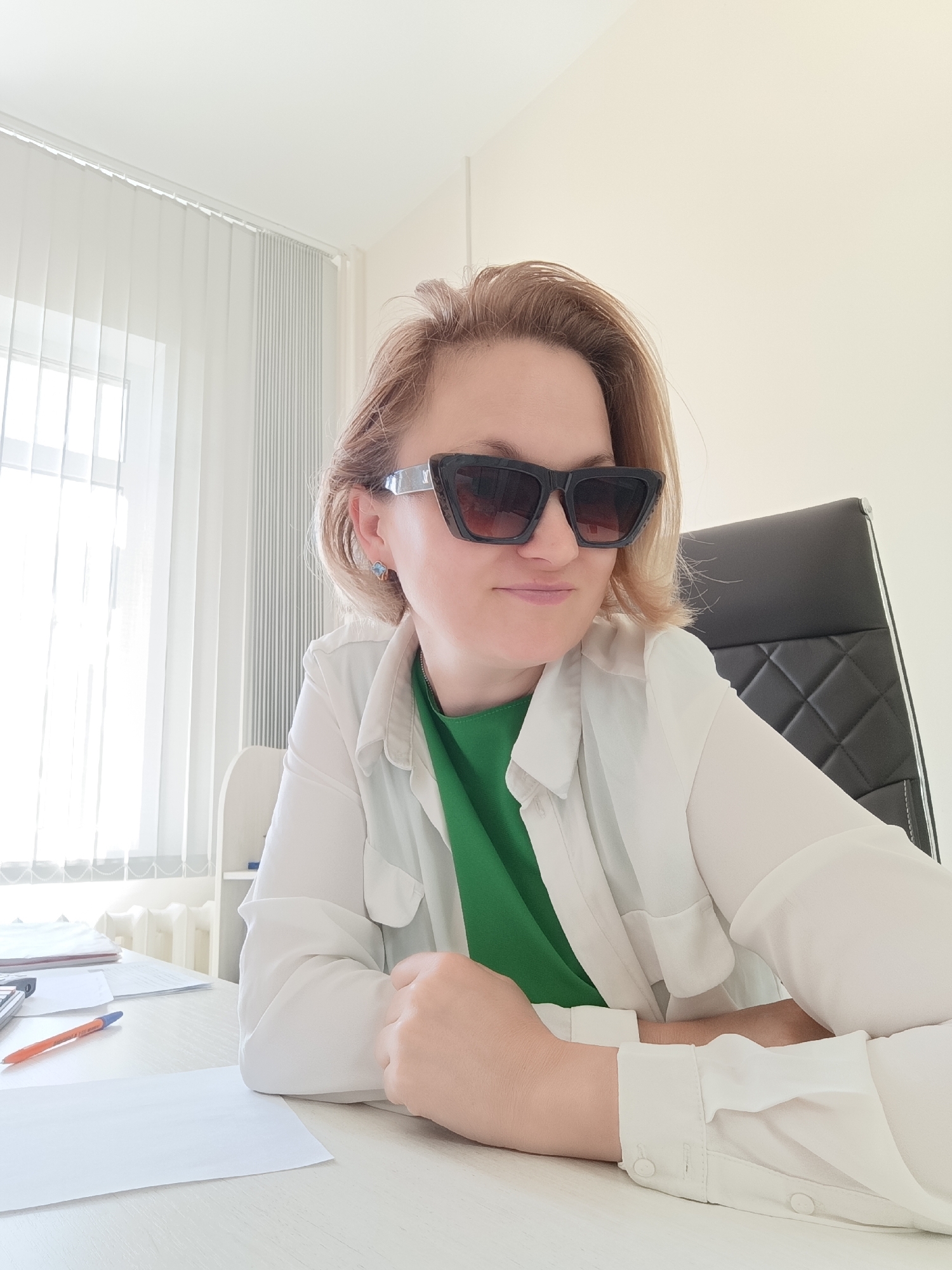 A blonde woman wearing sunglasses and a white shirt over a green top sits at a desk in an office. She is leaning forward, with her arms crossed and resting on the desk. There are papers and a pen on the desk. Behind her, a window with vertical blinds is visible, and to her right is a black office chair. The room appears to be a typical office setting. The image does not provide any specific location information within the city of Markovsky, Russia.