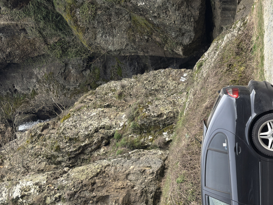 A dark-colored, four-door hatchback vehicle, consistent with a first-generation SEAT Leon model, is parked on a gravel shoulder in the lower-left foreground, its rear section partially visible. A subtle rear spoiler is present, and a light blue sticker adheres to the rear passenger window. The background is dominated by a large, rugged rock face covered in a mosaic of grey and green lichen, moss, and sparse vegetation, including bare tree branches and patches of dry, thorny bushes. A narrow waterfall cascades down the upper-left portion of this rock face. To the right, a deep, dark vertical crevice or gorge separates two massive rock formations, descending into shadow. A white marker, appearing to read "21 M," is affixed to the lower section of the rock face near the crevice. The ground immediately adjacent to the car and the base of the rock formation is characterized by loose gravel and scrub vegetation. The scene depicts a natural, mountainous environment, located in the vicinity of the city of Rila, Bulgaria. No individuals are visible.