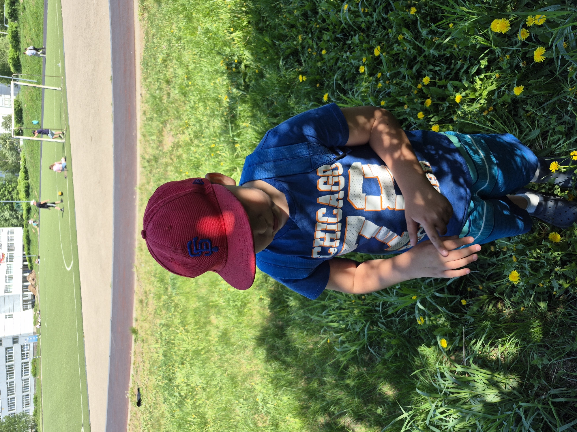 A young person wearing a red baseball cap and a blue Chicago jersey with the number 23 is lying in a field of grass and dandelions. The person's legs are bent, and they are looking down at their hands, which are resting on the grass. The location appears to be an outdoor area near a sports field with a running track. A building is visible in the background. Based on the context, it is likely the photo was taken in or near Chaikovsky, Russia.