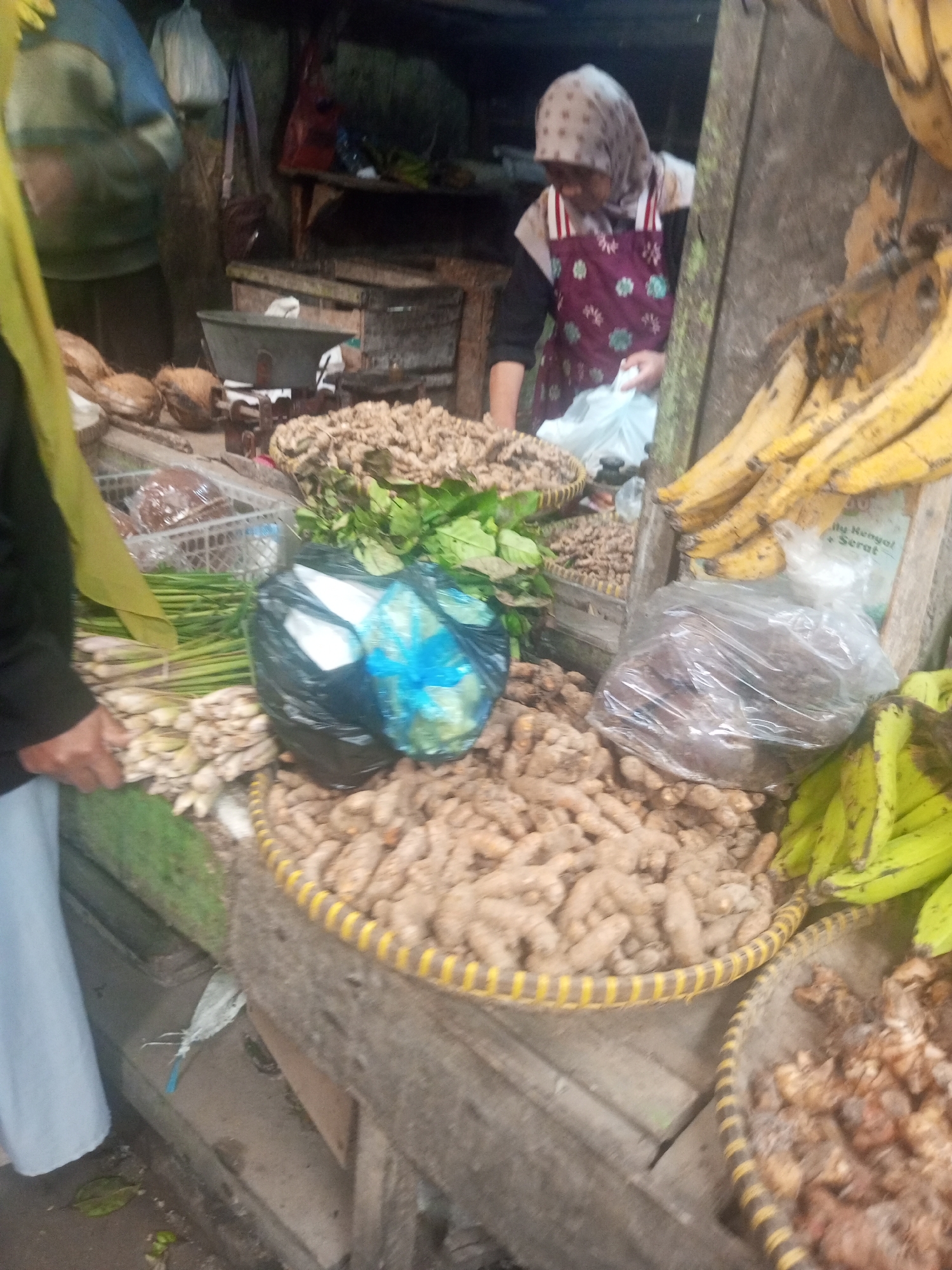 In Kalibeber, Indonesia, a marketplace scene unfolds, featuring a woman in a floral apron and headscarf attending to her stall. She is dressed in a dark shirt and white pants, with a maroon apron adorned with white and green floral patterns. In front of her are mounds of ginger and other root vegetables, arranged in woven baskets. One basket, prominently displayed in the foreground, is filled with a large quantity of ginger roots, its rim detailed with yellow and black stripes. To the right, a bunch of green bananas hangs, while to the left, more root vegetables, possibly lemongrass, are bundled together. A black plastic bag containing what appears to be white items is also visible. In the background, other market stalls are visible, with a scale and other produce suggesting a bustling trade. The overall impression is one of a lively local market.