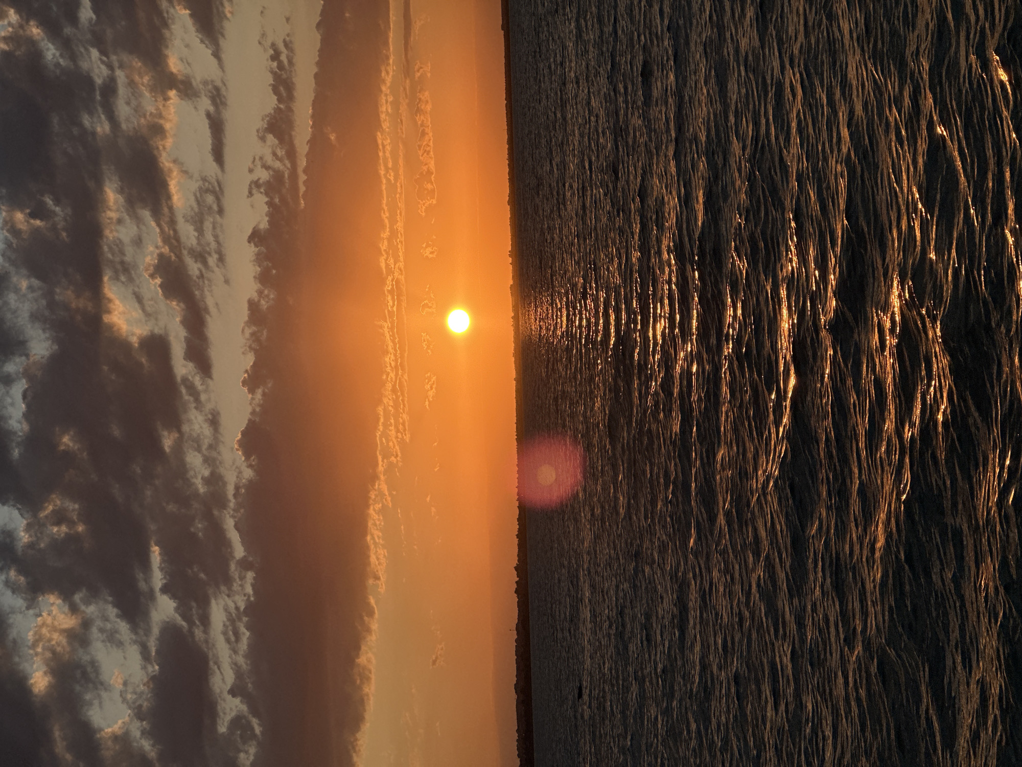 The image depicts a sunset over a body of water, possibly a lake or sea. The sun, a bright orange orb, is positioned slightly left of center on the horizon, casting a golden glow across the scene. The sky is filled with dramatic clouds, ranging in color from deep gray to warm orange, suggesting a dynamic weather pattern. The water surface is rippled, with sunlight reflecting off the waves in shimmering streaks of gold. In the distance, a faint silhouette of a treeline or buildings can be seen along the horizon. A red lens flare from the sun is visible in the middle right of the frame. The overall impression is one of natural beauty and tranquility. The image does not contain any identifiable landmarks or features that would indicate a specific location within the city of Kokshetau, Kazakhstan.