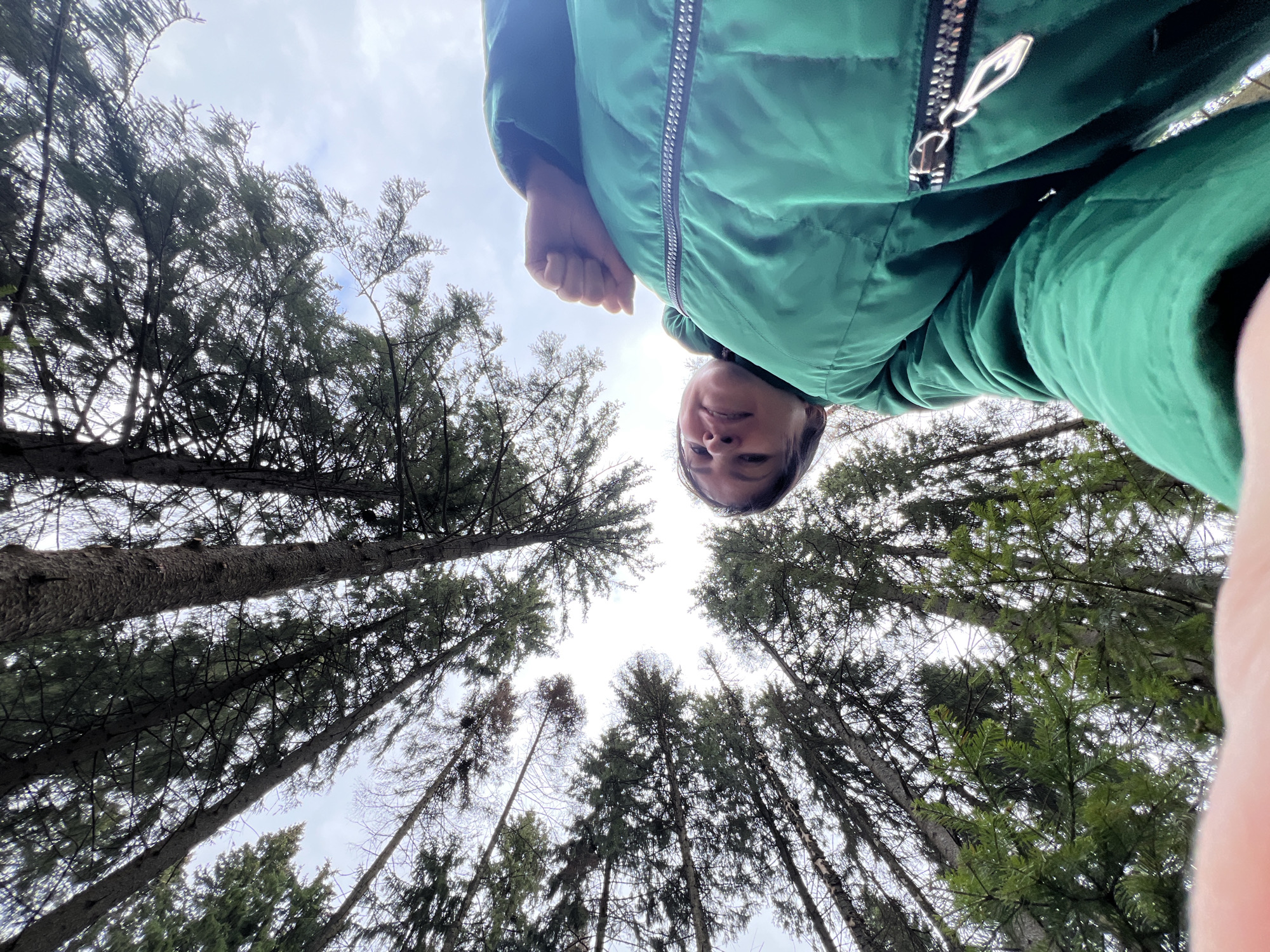 A low-angle shot looking up at a person wearing a green puffer jacket, with a background of tall trees reaching towards the sky. The sky is overcast. The person's face is visible and close to the camera. The location is likely a forest. Location Kosoturikha, Russia is not identifiable in this image.
