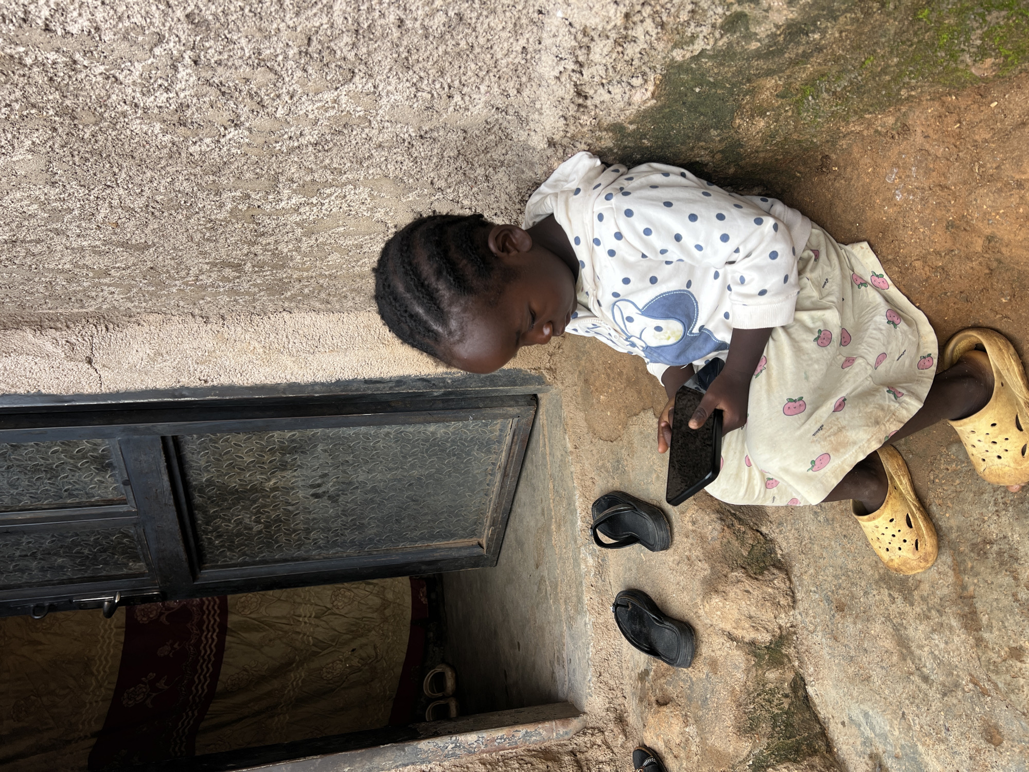 A young Black child sits on a stone step in front of a building, engrossed in a smartphone. The child is wearing a white hooded sweatshirt with navy blue polka dots and a blue cartoon character design, and a light-colored skirt patterned with pink strawberries. Yellow Croc-style shoes are on their feet. Two pairs of black flip-flops lie on the step to the child's left. The building wall behind the child is rough and textured, with patches of green moss. To the left, an open doorway reveals a glimpse of the interior, with a patterned curtain and what appear to be two pairs of white sandals on the floor. The overall scene suggests a simple outdoor setting. The location is identified as Jos, Nigeria.