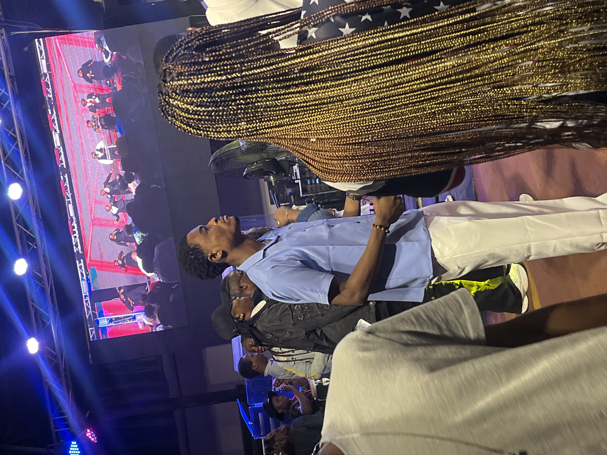 The image depicts a live music event or concert taking place in Ado, Nigeria. In the foreground, a person with long, braided golden-brown hair is the primary subject, obscuring the view of other individuals. To the left of this person is a man wearing a light blue collared shirt and white pants, facing to the right. Behind him, several other individuals are visible, some in casual attire, and one person is wearing a black baseball cap. In the background, a large LED screen displays a performance with multiple individuals on stage, dressed in dark clothing. The stage appears to be lit with colorful lights, including blue and white spotlights. The environment suggests an indoor or covered outdoor venue, with evidence of stage lighting and equipment. The crowd is gathered, indicating an audience observing the performance.