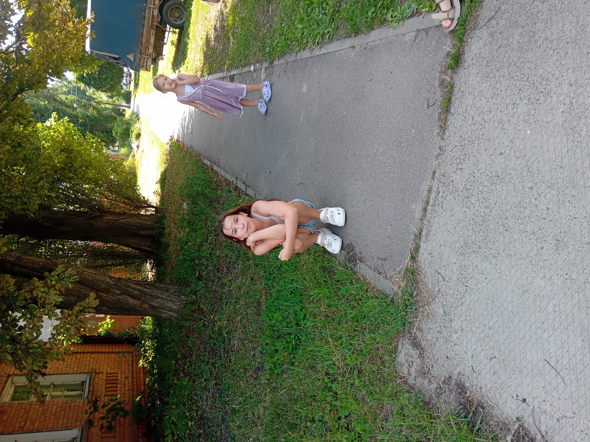 Two young girls are standing on a paved sidewalk next to a grassy area. The girl on the left is squatting on the sidewalk, facing the camera with her arms crossed. She is wearing a white tank top and blue shorts, and white Crocs. The girl on the right is standing further down the sidewalk, looking towards the camera. She is wearing a lavender dress and white Crocs. Behind the girls, there is a brick building, trees with green leaves, and a blue tarp-covered trailer. The scene appears to be outdoors in a residential area. The image is taken from a slightly elevated angle.