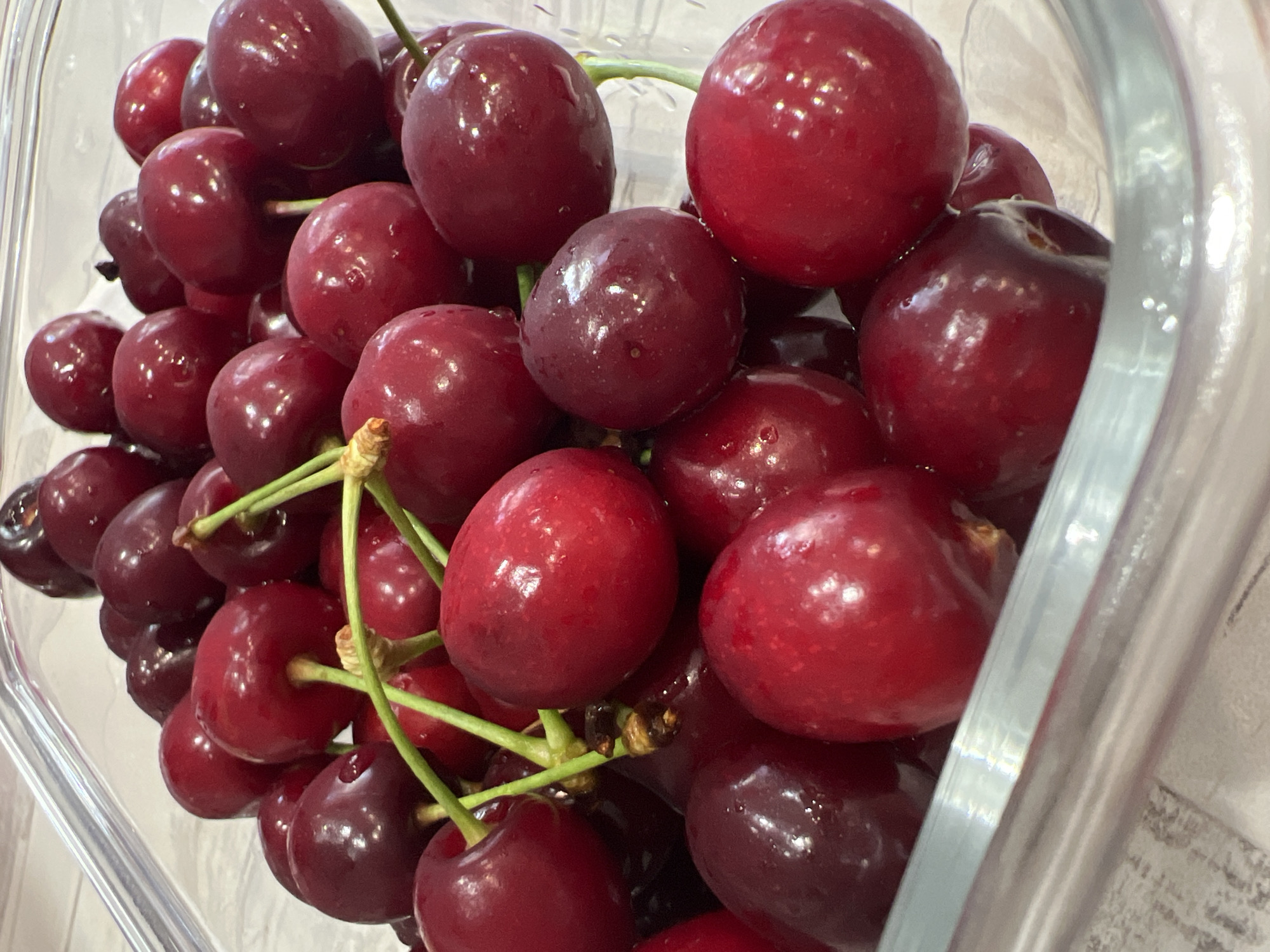 The location is Unknown, Unknown. The image displays a close-up view of numerous fresh red cherries contained within a transparent, rectangular dish or container. The cherries vary in color from bright crimson to dark burgundy, with many exhibiting small, reflective water droplets on their glossy surfaces. Several green stems are visible, some branched and connecting multiple cherries, while others appear as single attachments. The container is almost completely filled with the cherries. The background consists of a light-colored, subtly patterned surface, likely a countertop, which is partially visible around the edges of the container and appears slightly out of focus.