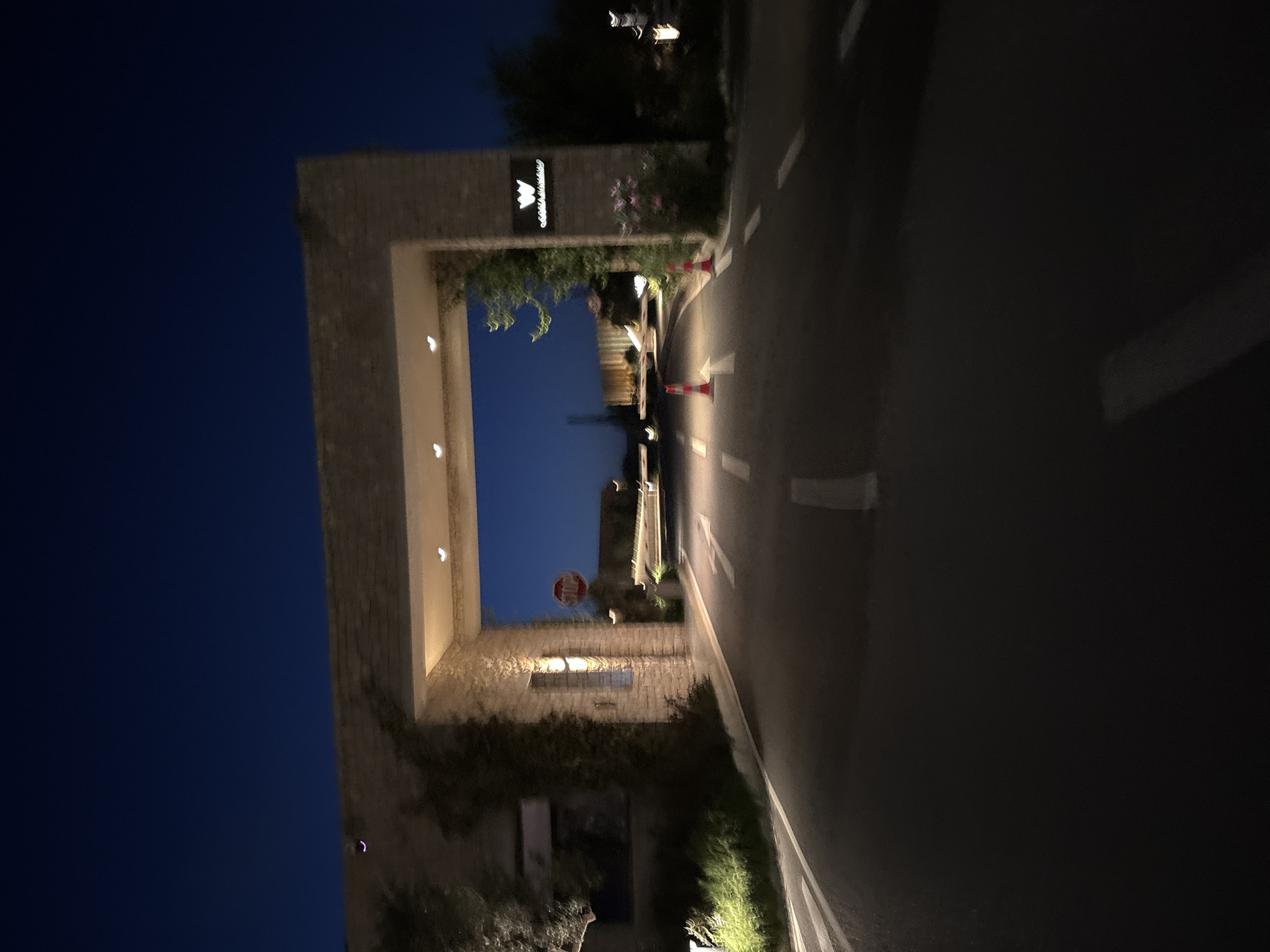 The image captures a nighttime view of a stone archway serving as an entrance, likely to a property or resort, with a road leading through it. The perspective is from the side of the road, looking towards and slightly through the archway.
The archway is constructed from light-colored stone or masonry, with subtle recessed lights illuminating its inner passage. Above the archway, on the right side, an illuminated sign is visible, displaying a crown-like logo and the text "Costa Navarino." Green foliage and bushes grow around the base and sides of the archway.
The asphalt road in the foreground features white dashed lines and white directional arrows painted on its surface. Several orange traffic cones are placed along the right side of the road, guiding traffic towards the archway. A red octagonal "STOP" sign is visible further down the road, beyond the archway, though it is slightly out of focus.
The sky is a deep, dark blue, indicating the scene takes place at night. Artificial lighting illuminates the archway, the sign, and parts of the road and surrounding vegetation. No people or vehicles are present, creating a quiet and serene atmosphere. The scene suggests a formal and well-maintained entrance, consistent with a resort or significant establishment in Pylos, Greece.