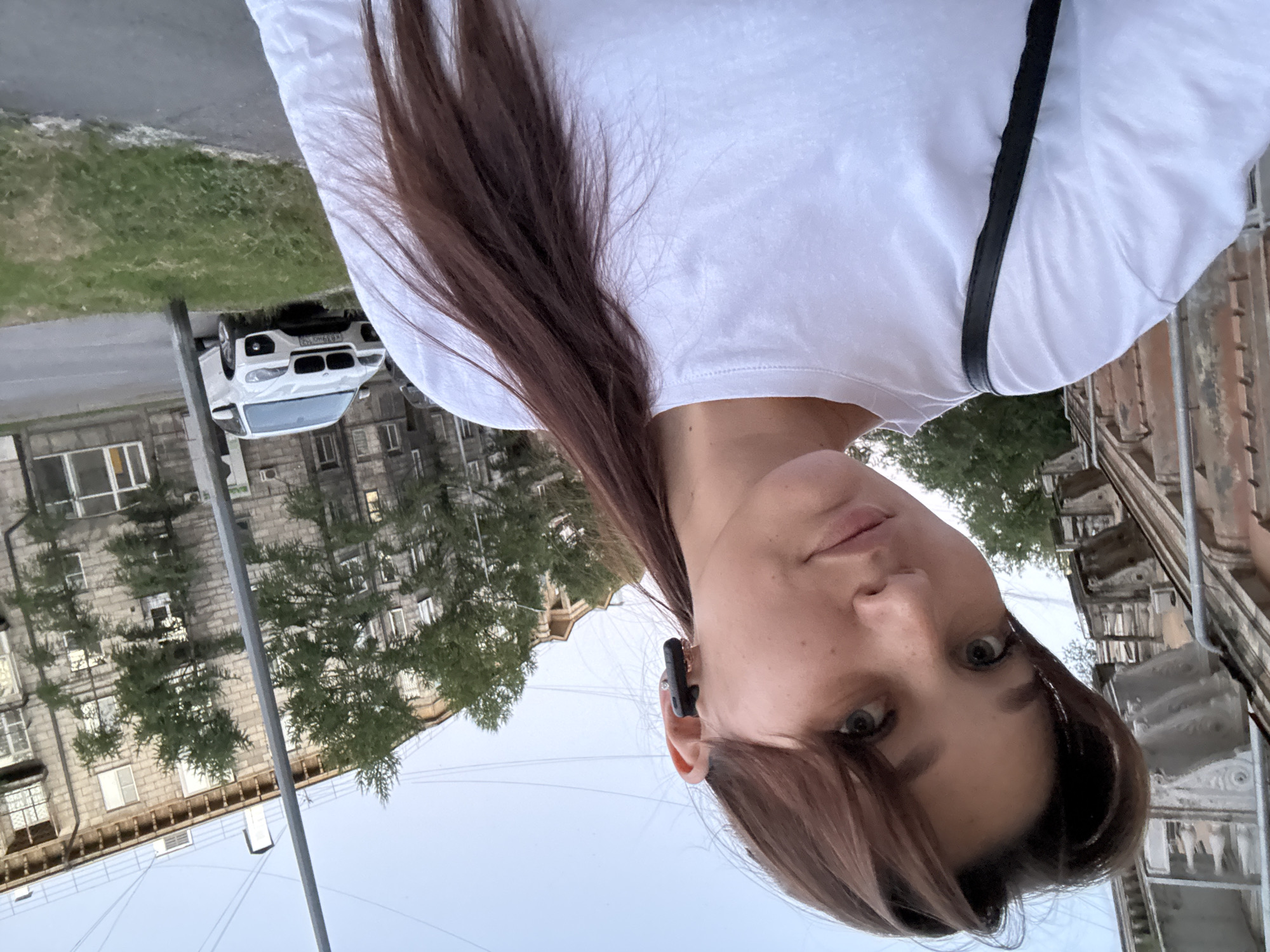 In Novokuznetsk, Russia, a young woman with brown hair pulled into a ponytail takes a selfie on a sidewalk in front of an apartment building. She is wearing a white t-shirt and black earbuds. A white BMW is parked on the street behind her, and trees line the sidewalk. The sky is overcast, and the overall atmosphere appears to be casual and urban.
