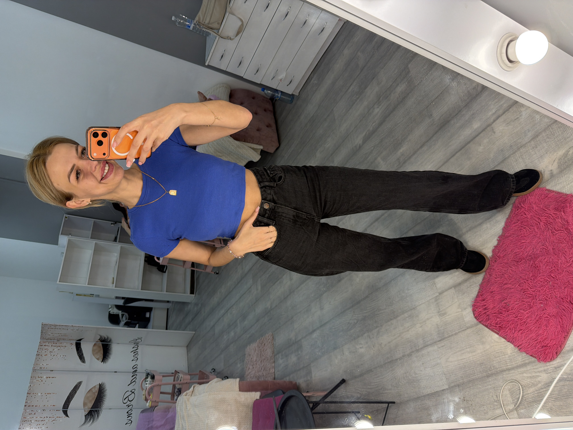 A blonde woman stands in a room with gray wood-look flooring, taking a mirror selfie with her orange-cased smartphone. She is wearing a royal blue cropped t-shirt, black high-waisted jeans, and black shoes. She has a gold necklace with a rectangular pendant and a thin gold bracelet on her left wrist. Her right hand is in her jeans pocket, and her left hand holds the phone. To her left is a white folding screen with a large graphic of eyelashes. Behind her are shelves, a pink trolley, and a dresser with several drawers. A bright pink shag rug is visible on the floor in front of her. The scene appears to be a personal beauty studio or salon. The image does not contain enough information to determine the specific location in Kyiv, Ukraine.