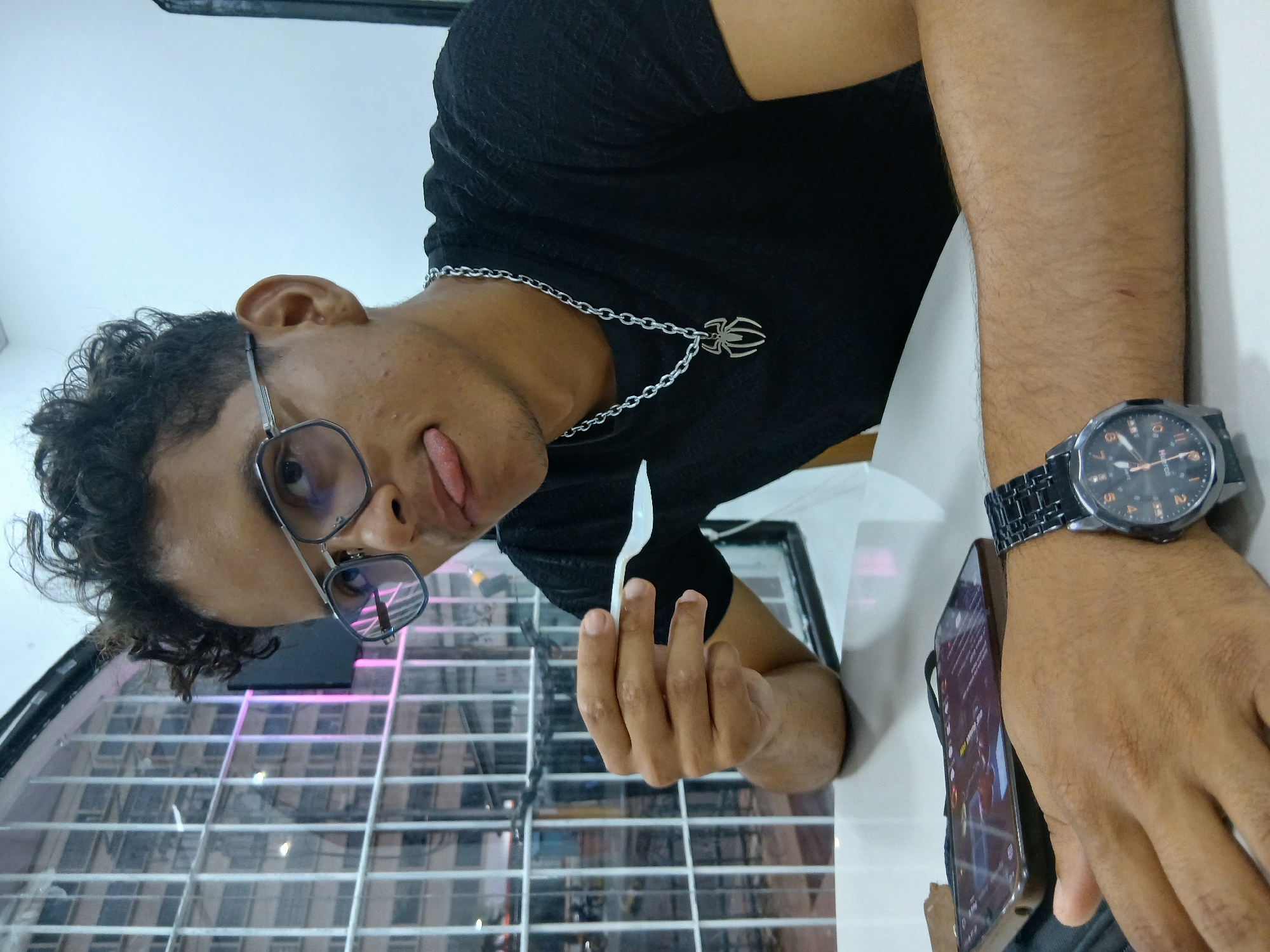 An upward-angled, close-up photograph captures a young person, likely indoors in Juan Griego, Venezuela. The individual has brown skin, dark curly hair, and wears dark-rimmed glasses. They are dressed in a black short-sleeved t-shirt and accessorized with a silver chain featuring a spider-shaped pendant around their neck. A dark-faced wristwatch with a metallic band, clearly showing the "FOSSIL" brand on its face, adorns their left wrist.
The person is looking towards the camera with a playful expression, sticking out their tongue. Their right hand casually holds a white plastic spoon. Next to their left arm, a metallic-backed smartphone is visible, its screen displaying blurred content that is unreadable. The background above the person is a plain, light-colored wall or ceiling. Below them, through large windows or glass panels, a grid-like structure and reflections of distant buildings or urban elements are discernible, illuminated by what appears to be daylight. Some pinkish-purple artificial lights are also visible in the reflected background. The scene conveys a casual and lighthearted mood.