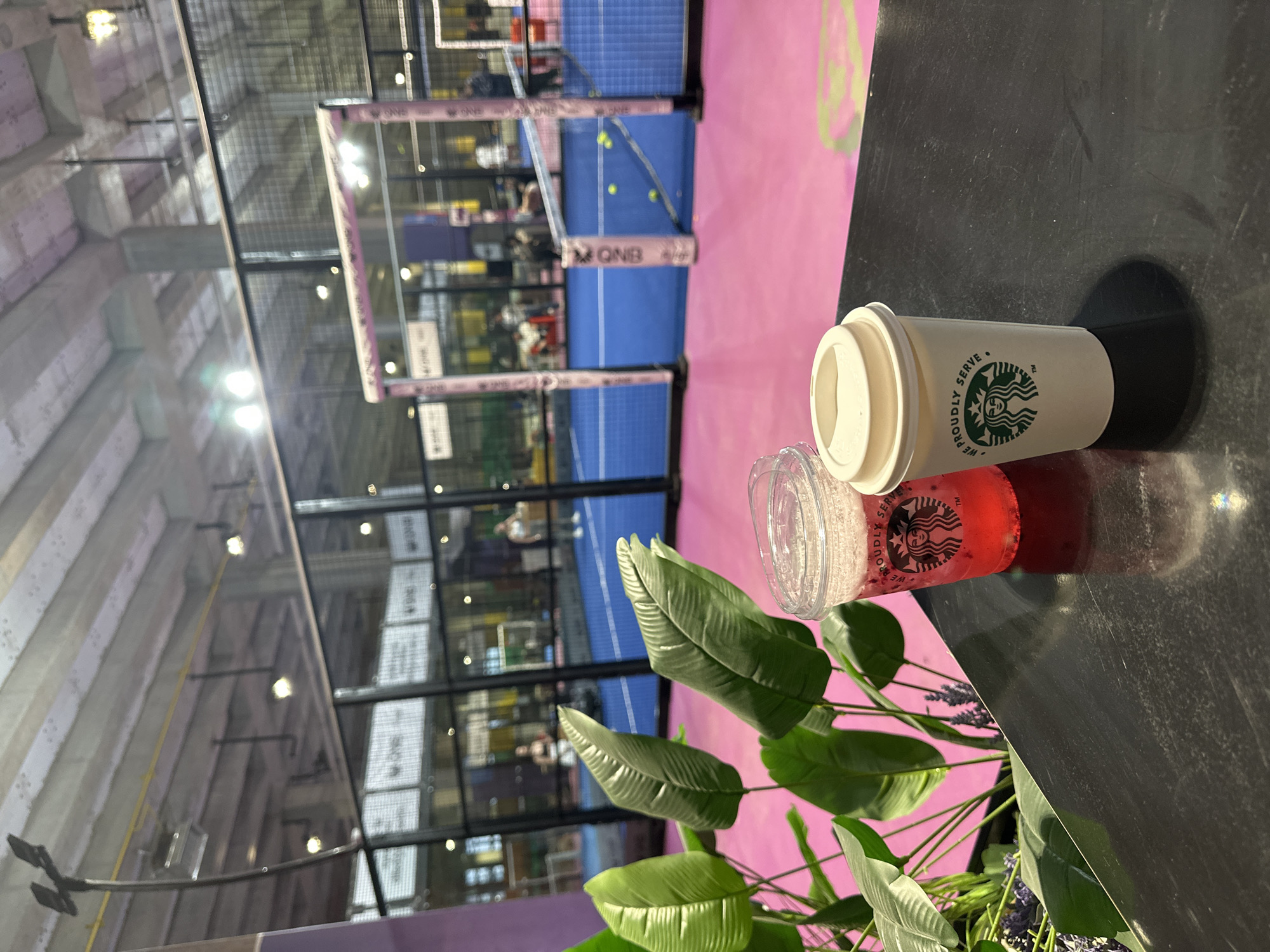 The image shows a Starbucks paper cup and a clear plastic Starbucks cup filled with a red liquid placed on a dark countertop. Green plants are also present on the countertop. In the background, there is a padel tennis court with blue and pink surfaces enclosed in a black metal frame. People are visible playing or spectating on the court. Given the presence of padel tennis courts, the location could be a sports complex or a venue that hosts such facilities in Esenyurt, Turkey.