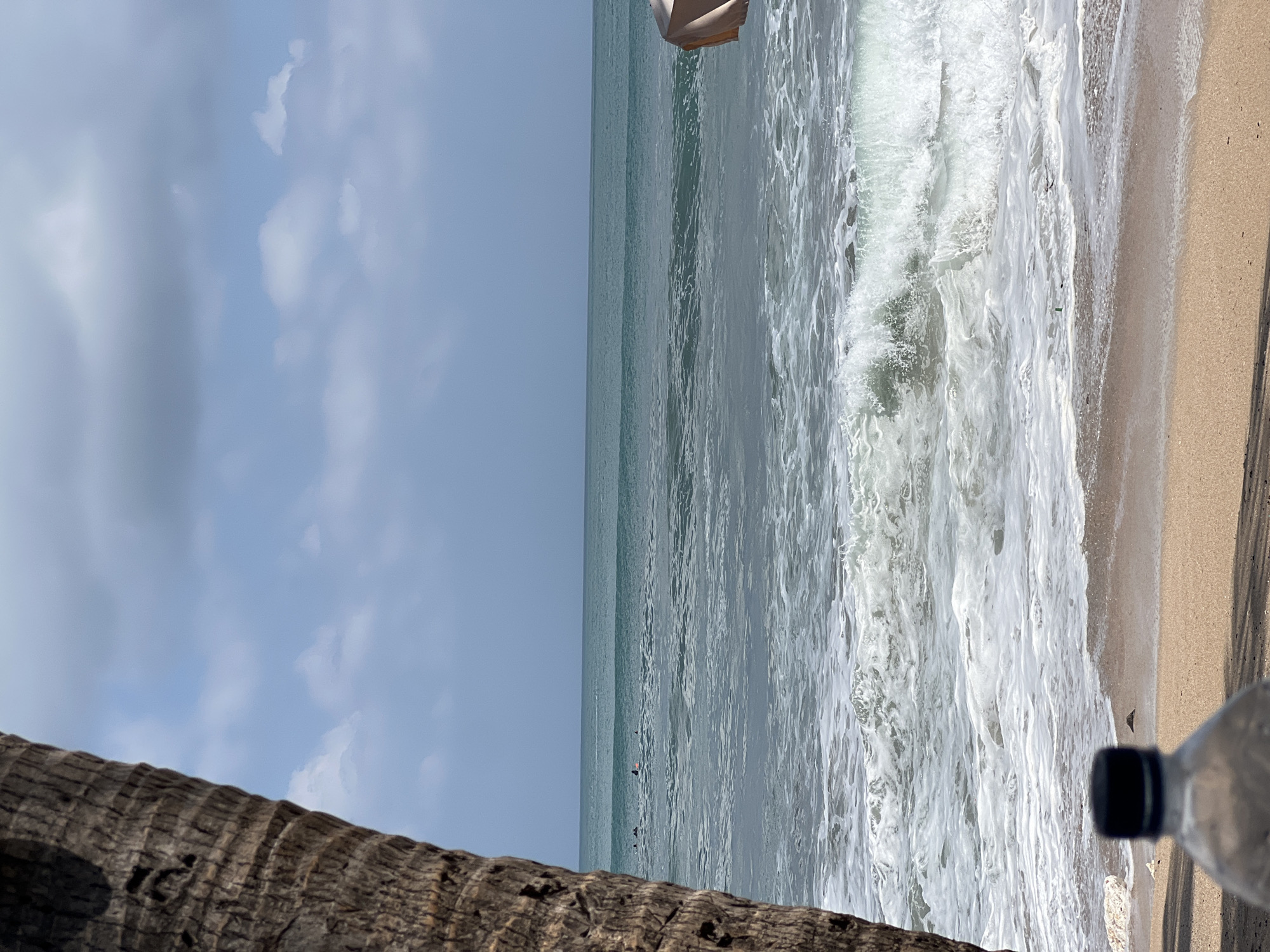 The image depicts a beach scene in Kuta, Indonesia, featuring the ocean, a sandy shore, and a portion of a palm tree trunk in the foreground on the left. The ocean exhibits waves breaking near the shore, with calmer water further out where several individuals are visible, some appearing to be surfing. The sky is partly cloudy with a light blue hue. In the bottom foreground, the top of a plastic bottle with a black cap is visible, partially obscuring the sand. To the right, the edge of a light-colored beach umbrella is present. The overall environment suggests a relaxed beach setting under a bright sky.