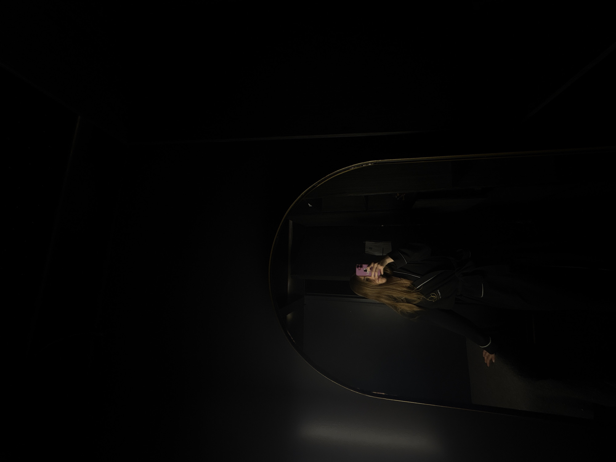 The image shows a reflection in a round mirror within a dark, possibly indoor, setting. The reflection reveals a person holding a pink mobile phone in front of their face. The person has long, blonde hair and is wearing a dark-colored outfit with white trim. The environment is largely undefined due to the darkness, but the mirror's surroundings appear to be dark walls. The city of Ciudad López Mateos, Mexico is mentioned in the prompt, but there is no information in the image to confirm that this is where the image was taken.